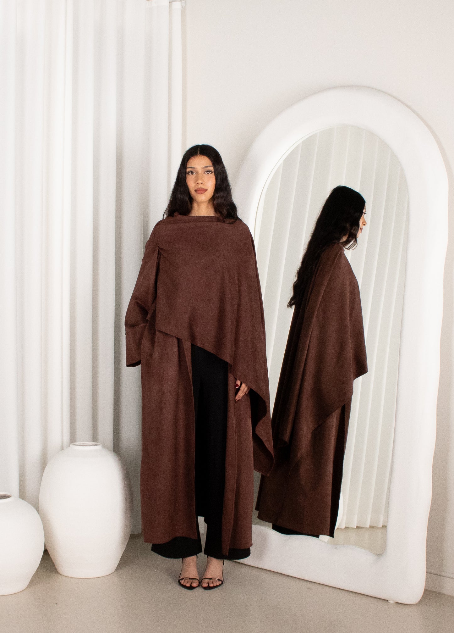 Woman wearing a brown poncho standing in front of a mirror in a minimalistic setting.