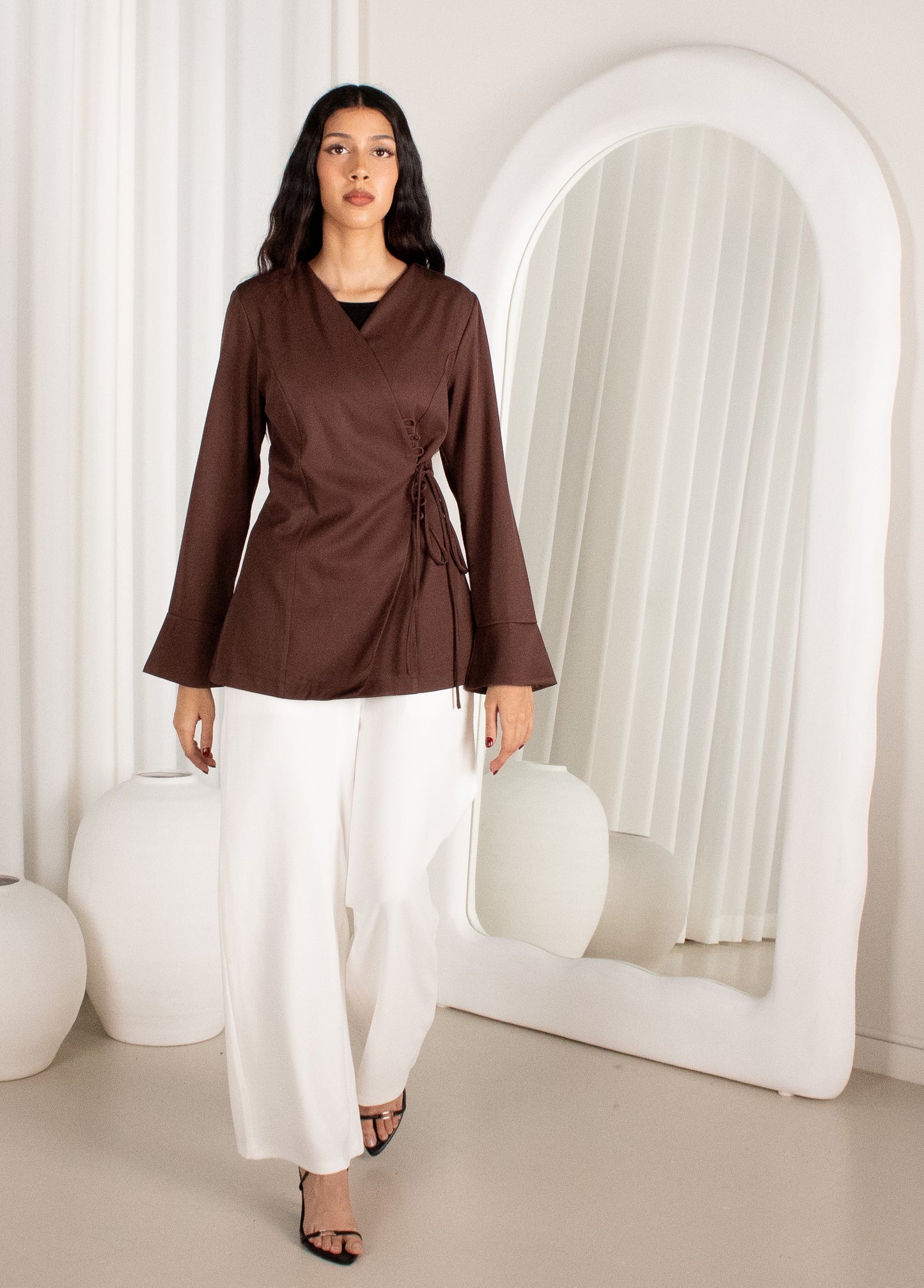Woman wearing a brown top and white pants standing in a minimalistic room with a large mirror.