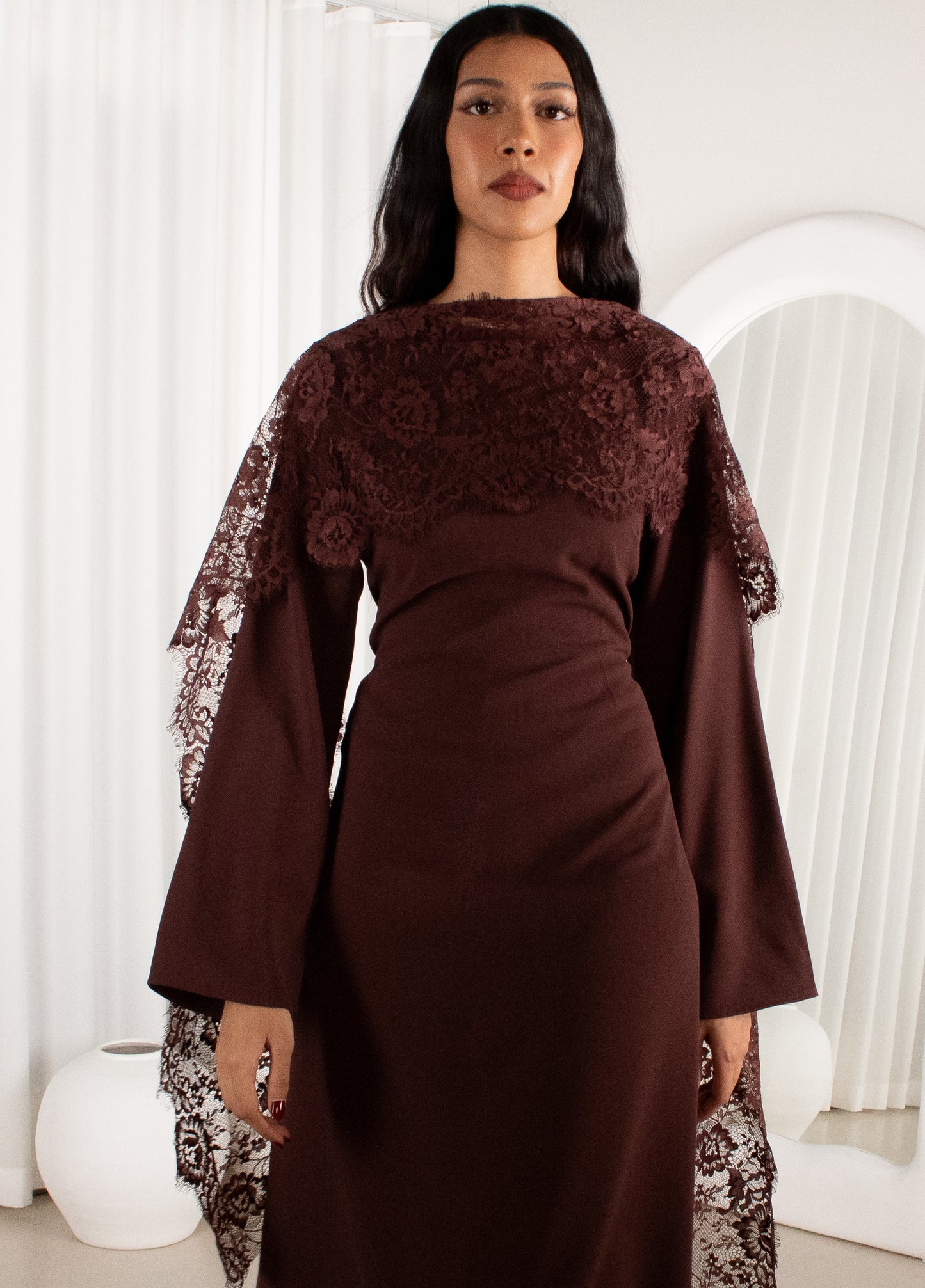 Woman wearing a brown dress with lace details in a white room.