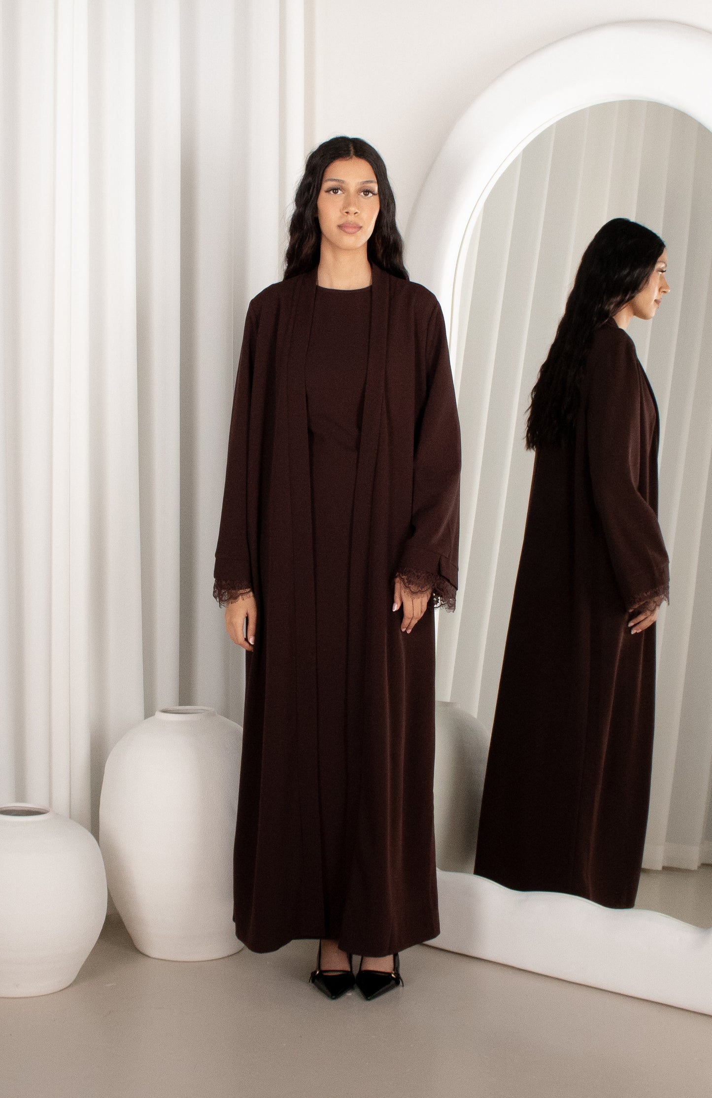 Woman wearing a brown abaya in front of a mirror with white curtains.