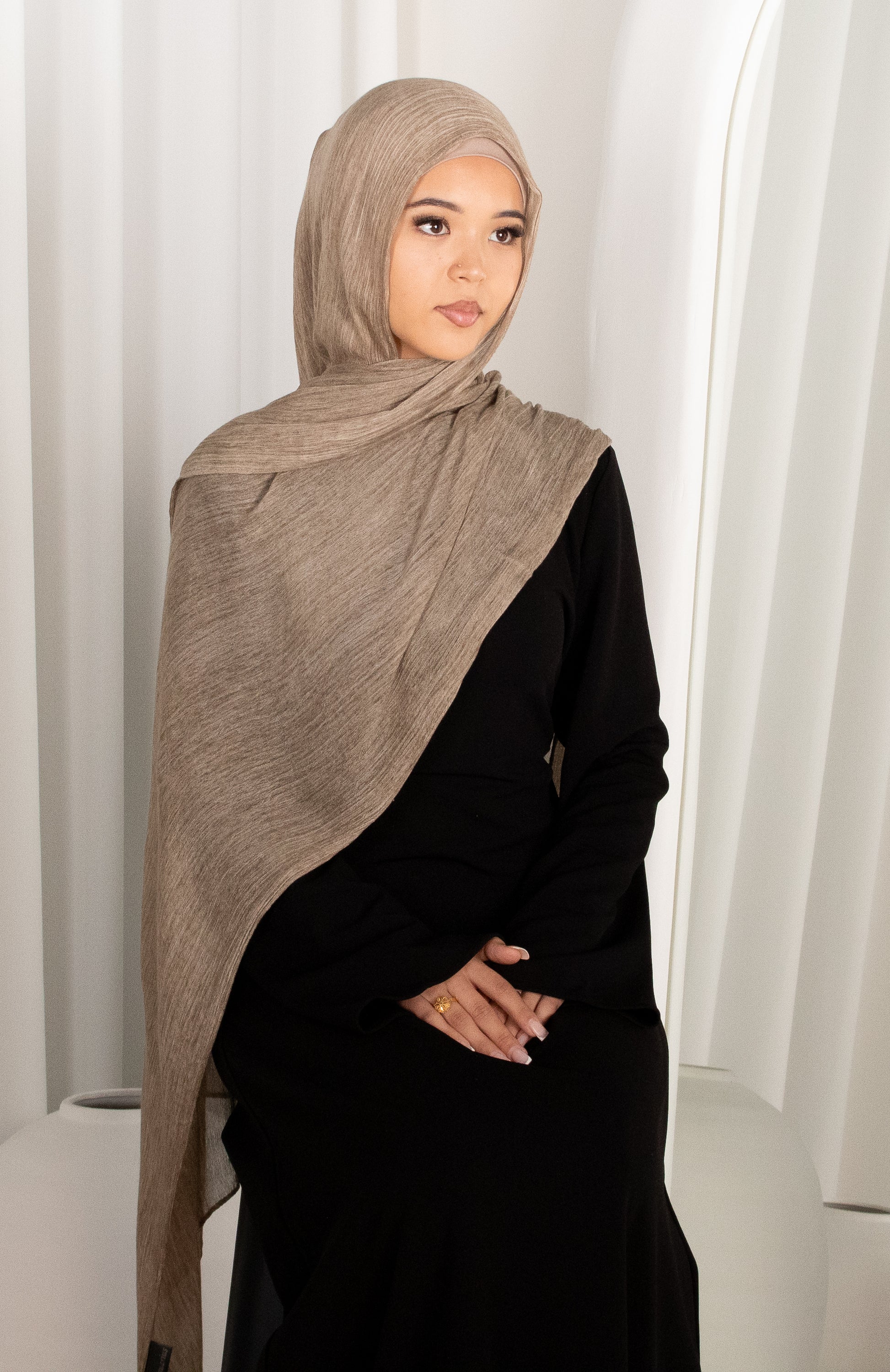 Woman wearing a beige hijab and black outfit against a white curtain background