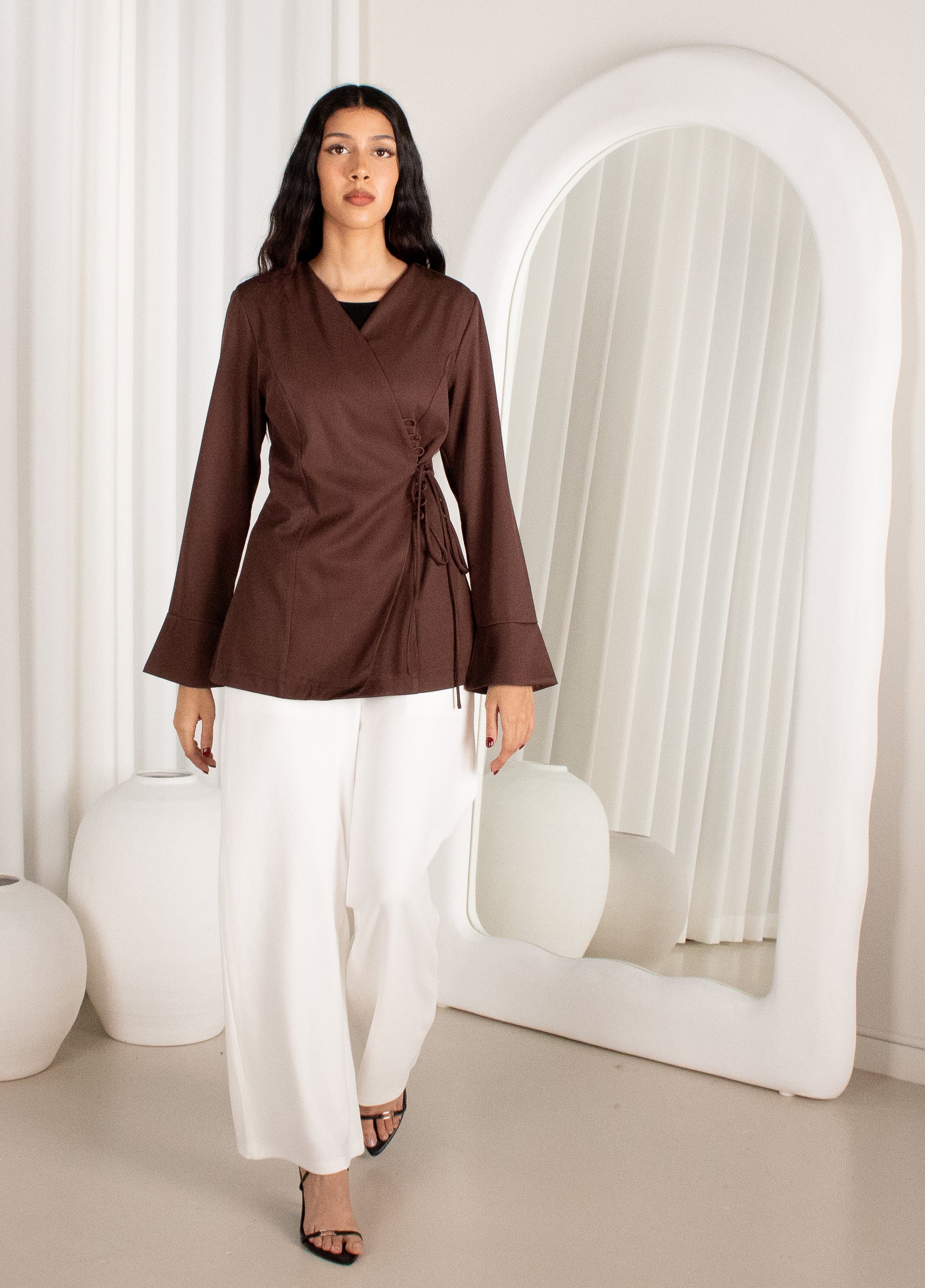 Woman wearing a brown top and white pants standing in a minimalistic room with a large mirror.