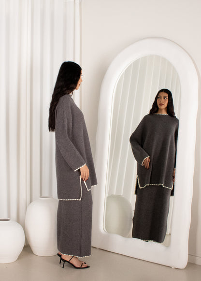 Woman in a gray outfit standing in front of a large mirror in a minimalistic room.