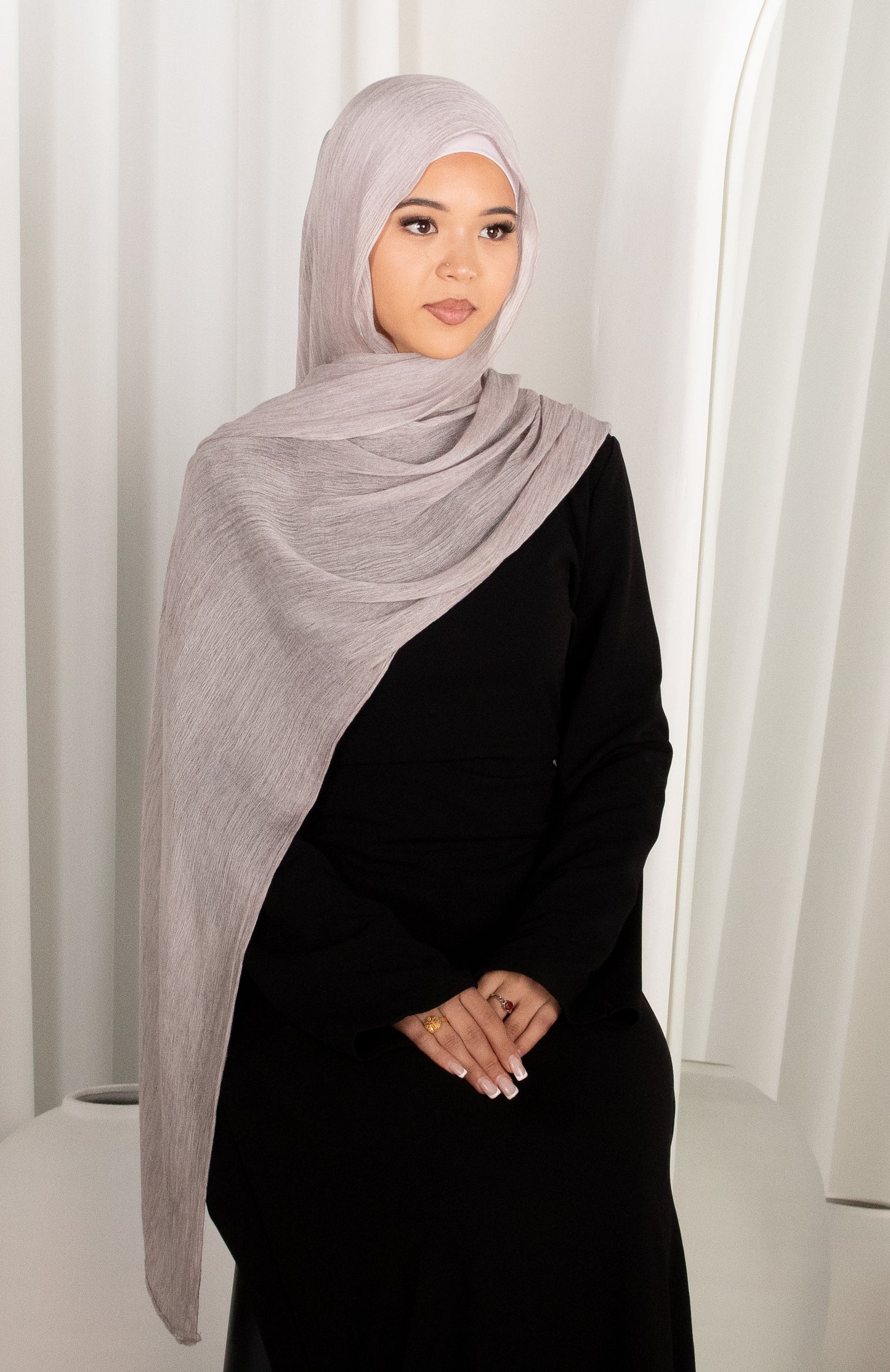 Woman wearing a gray hijab and black outfit against a white curtain background