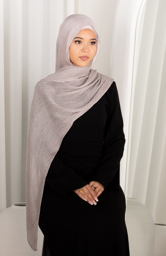 Woman wearing a gray hijab and black outfit against a white curtain background