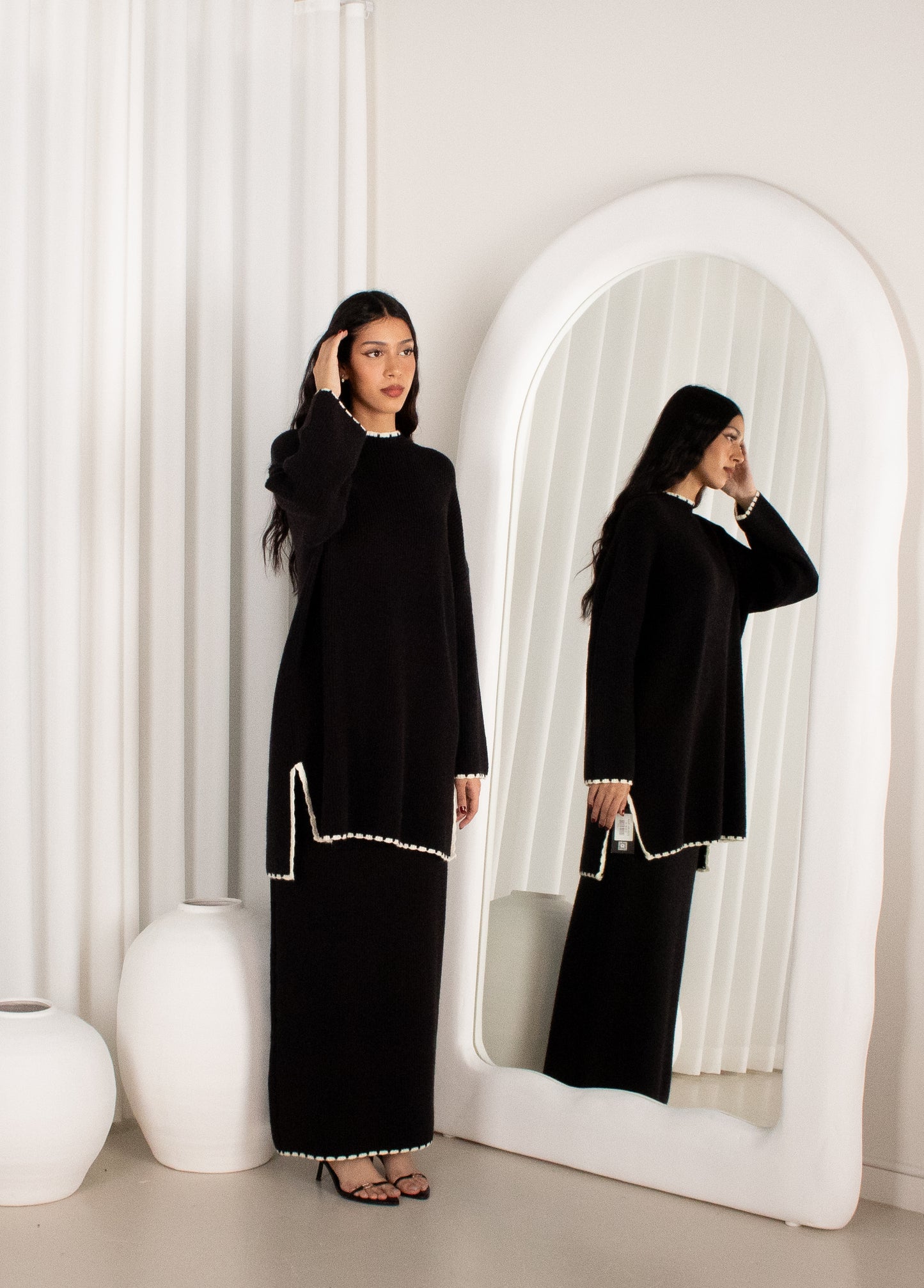 Woman in a black dress standing in front of a mirror in a minimalistic room.