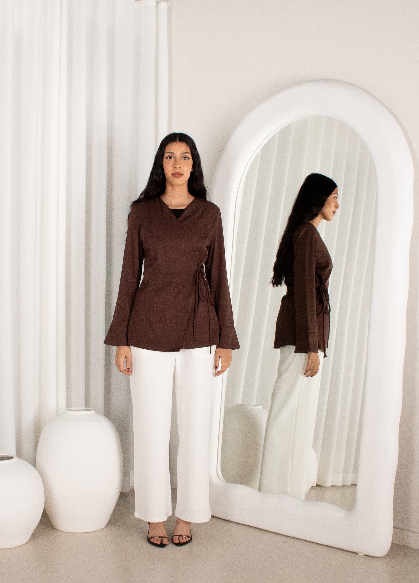 Woman in a brown top and white pants standing in front of a mirror in a minimalistic room.