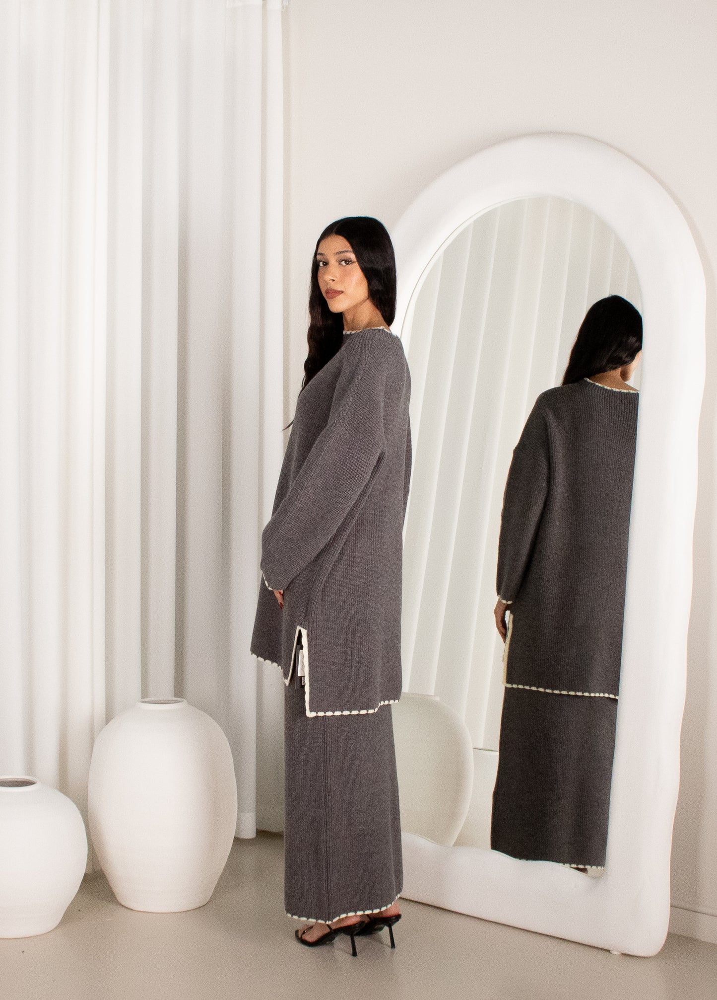 Woman in a gray dress standing in front of a white mirror in a minimalistic room.