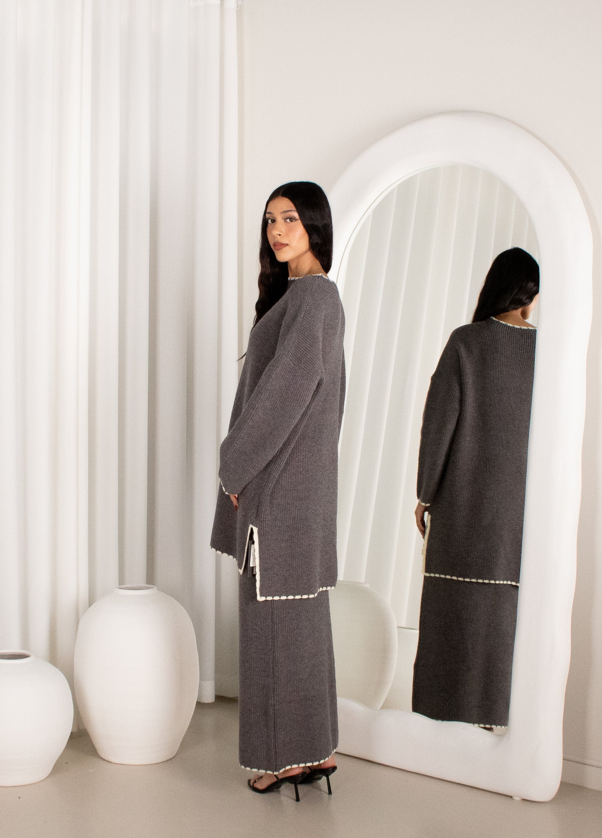 Woman in a gray dress standing in front of a white mirror in a minimalistic room.