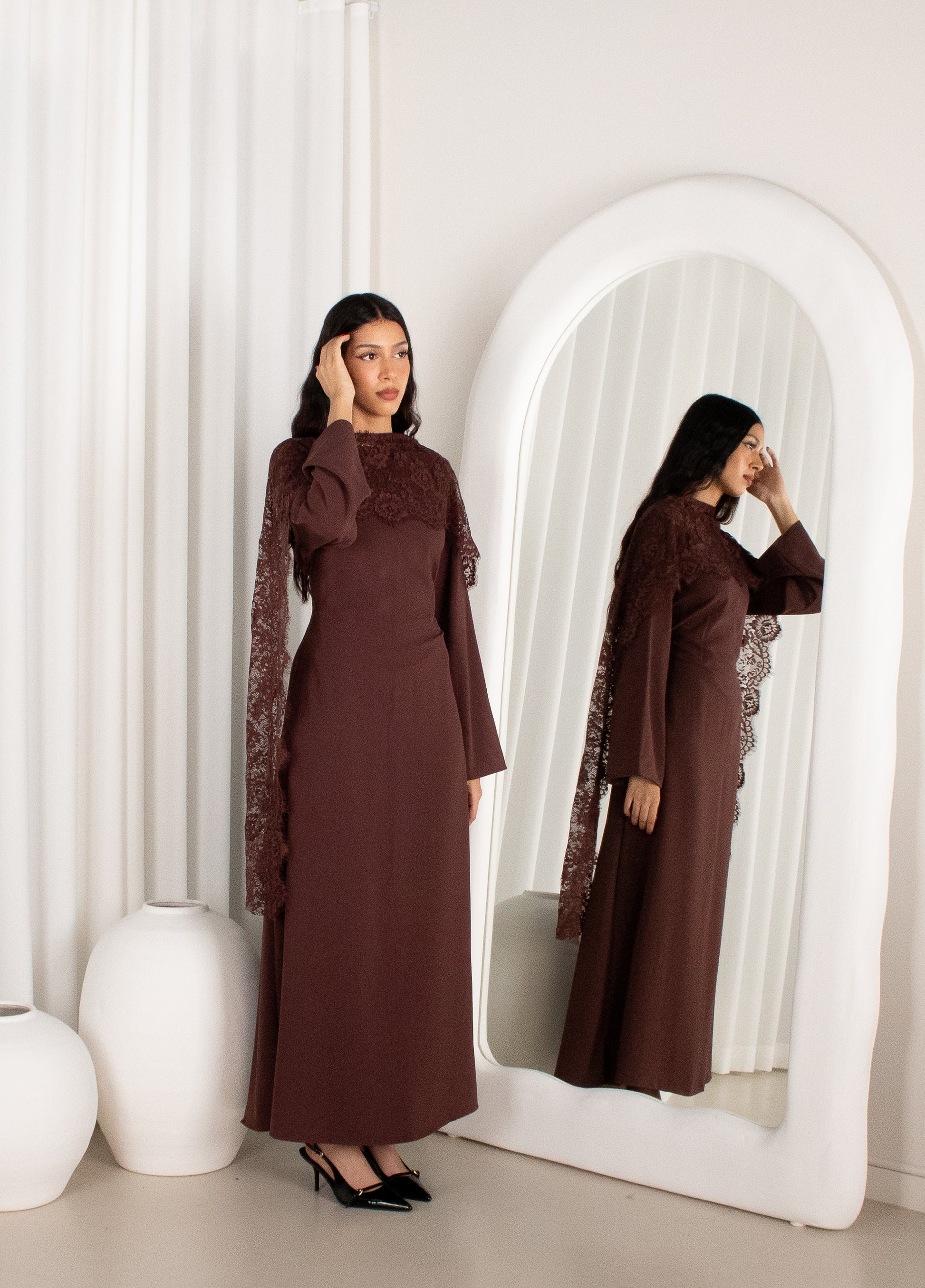 Woman in a brown dress standing in front of a mirror in a minimalistic setting.