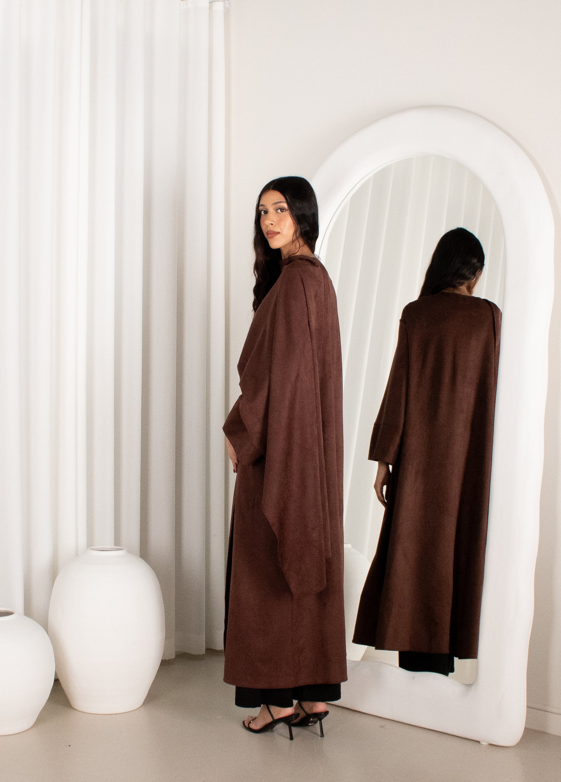 Woman wearing a brown coat standing in front of a white curtain and mirror.