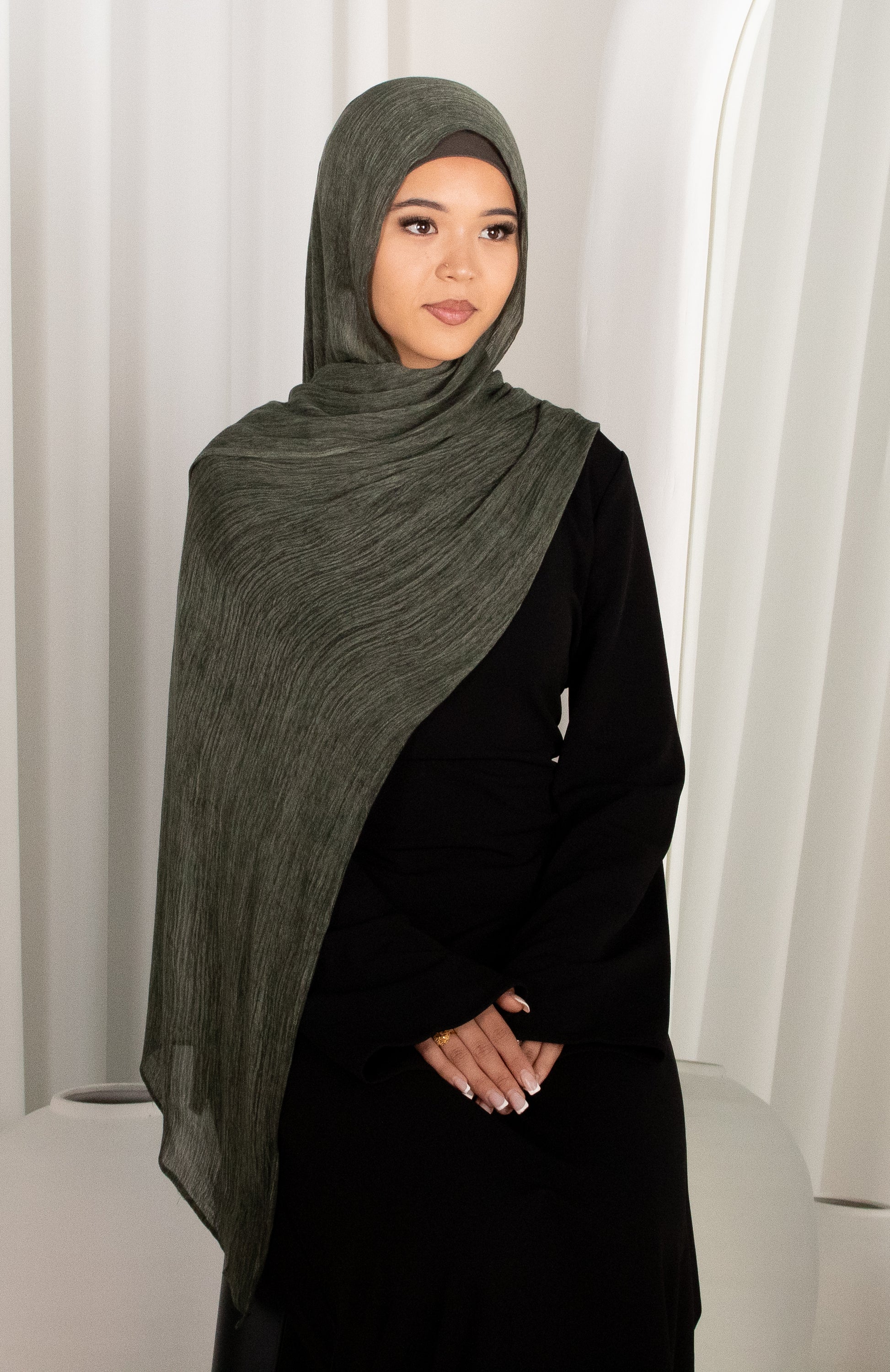 Woman wearing a dark green hijab and black outfit against a white curtain background