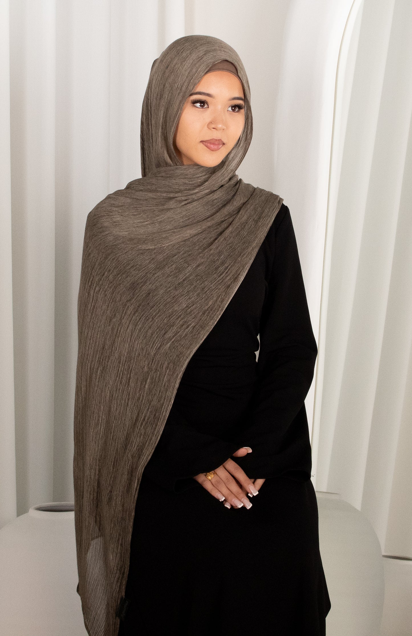 Woman wearing a brown hijab and black outfit against a white curtain background