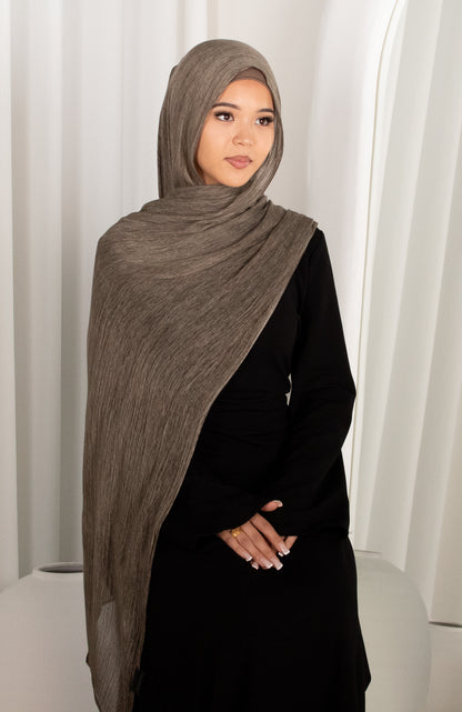 Woman wearing a brown hijab and black outfit against a white curtain background
