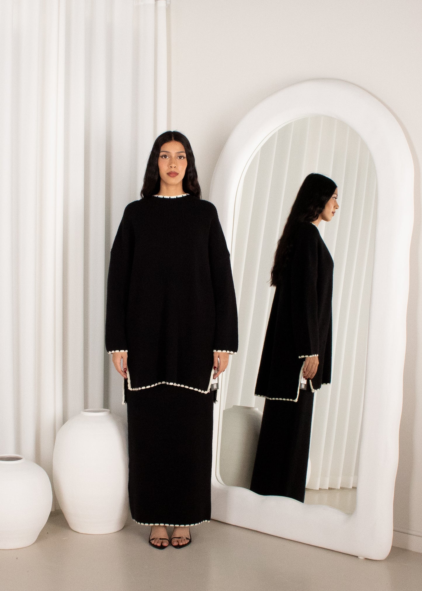 Woman in a black outfit standing in front of a mirror with white curtains.
