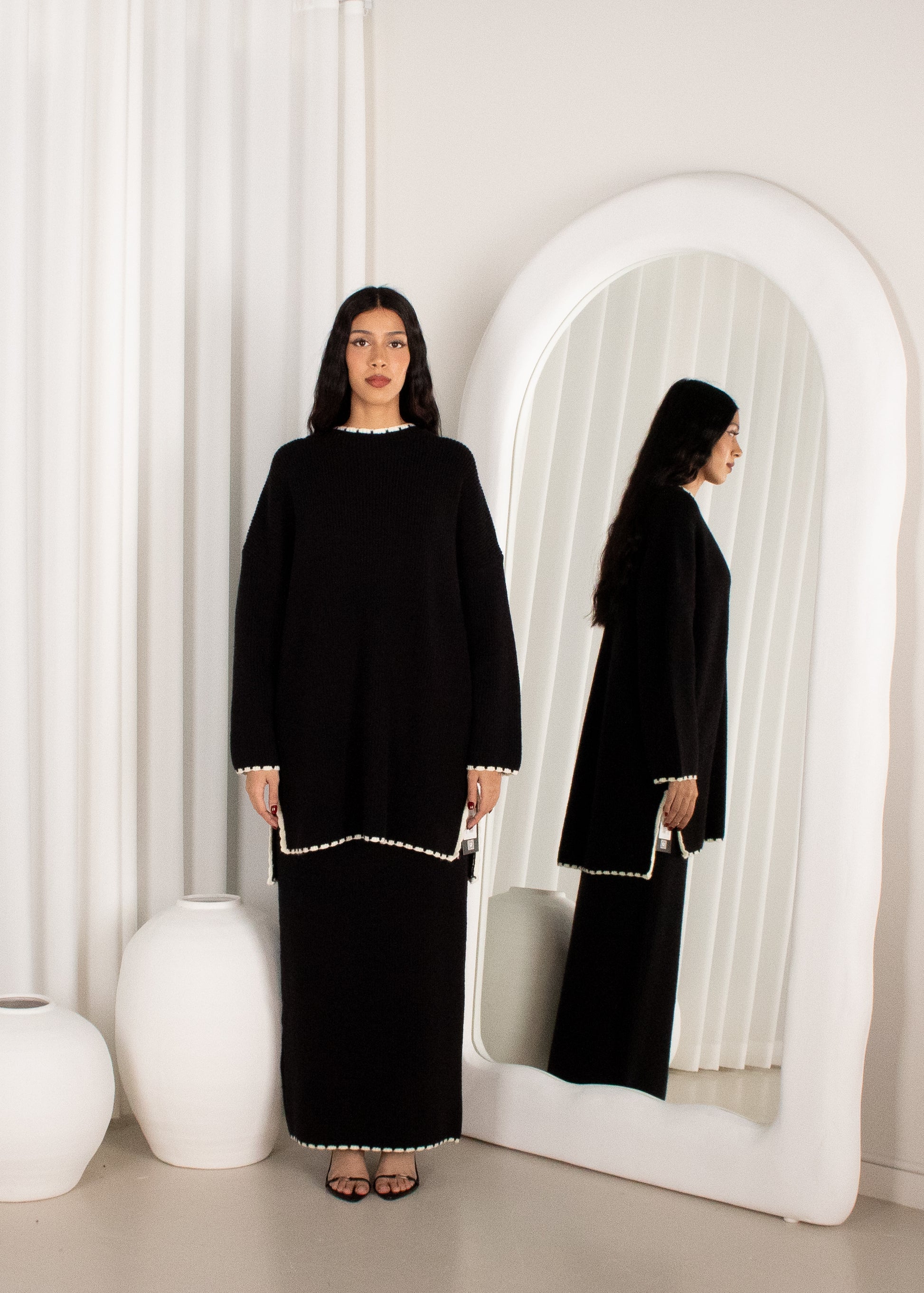 Woman in a black outfit standing in front of a mirror with white curtains.