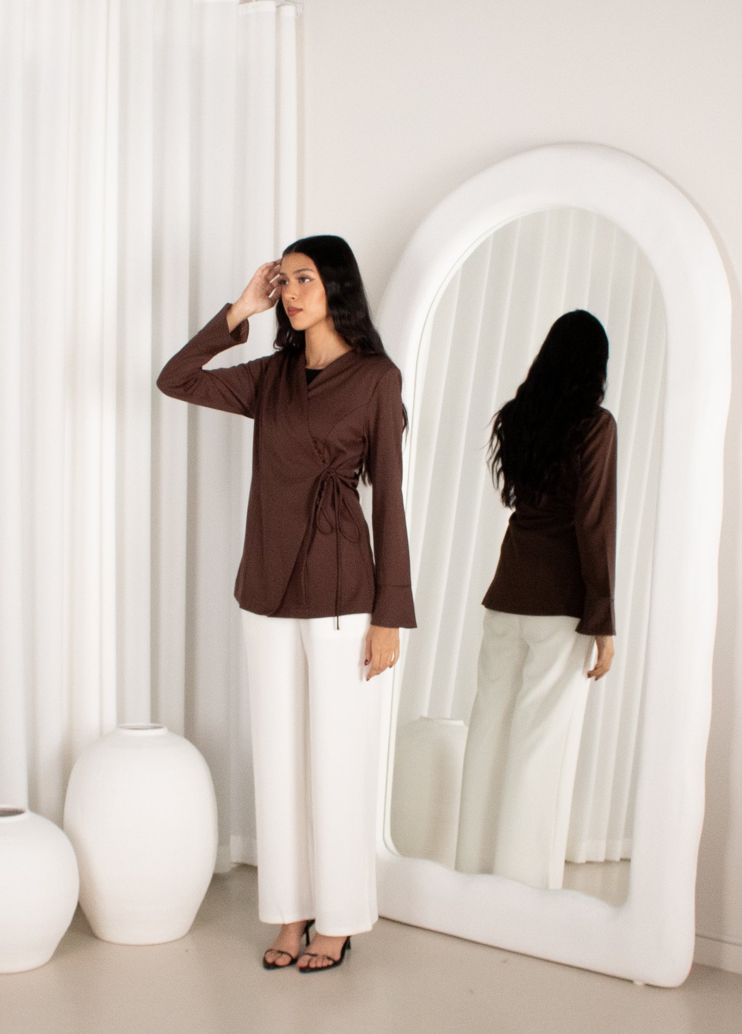 Woman in a brown top and white pants standing in front of a large mirror.