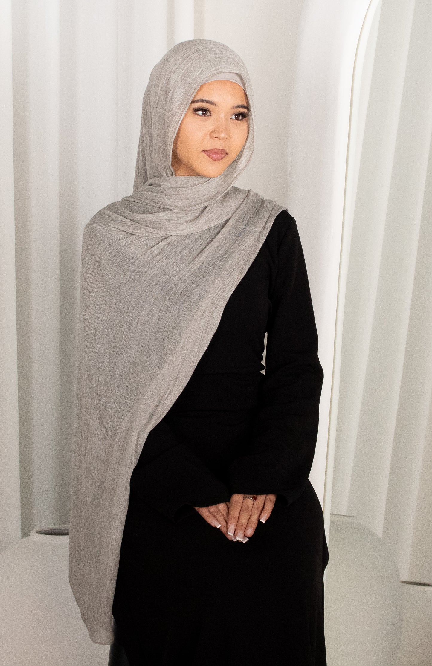 Woman wearing a gray hijab and black outfit against a white curtain background