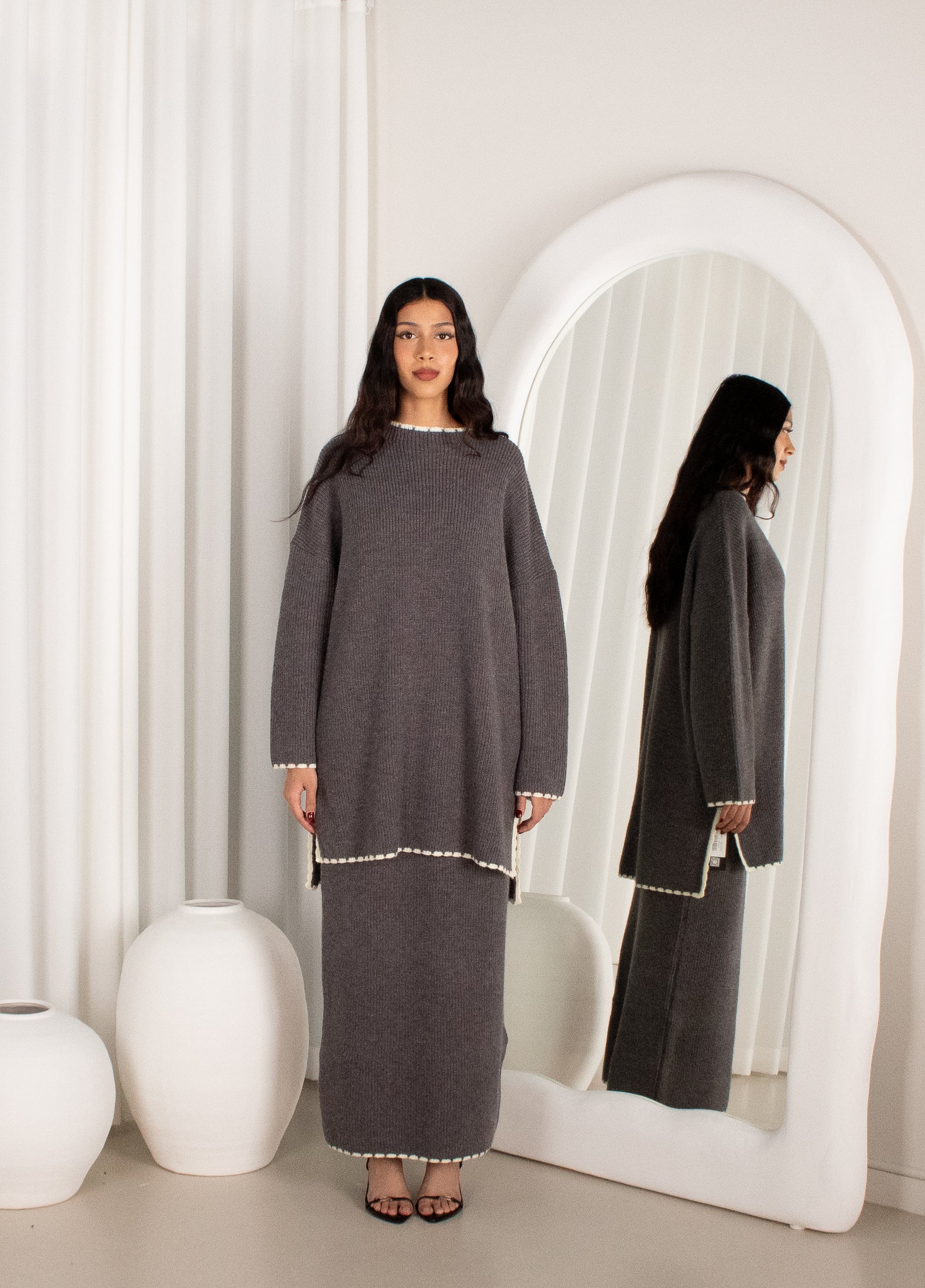 Woman wearing a gray dress standing in front of a mirror in a minimalistic room.