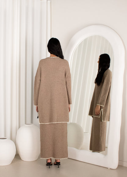 Woman in a long brown dress standing in front of a mirror in a minimalistic room.