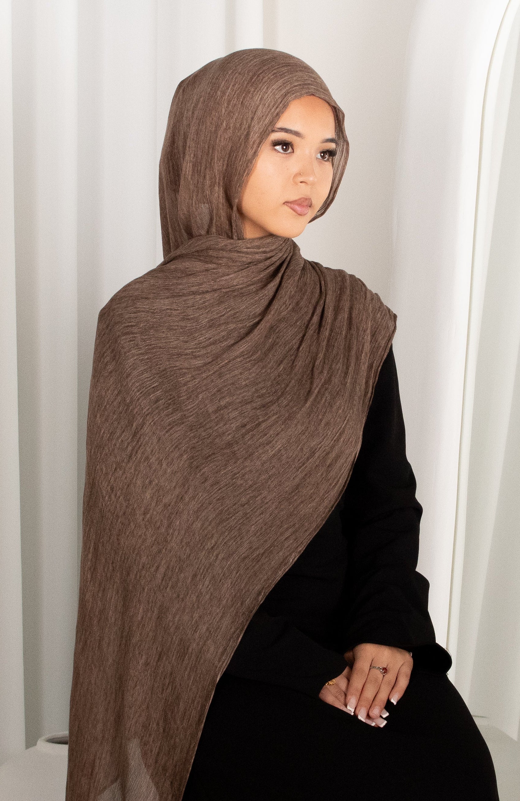 Person wearing a brown hijab and black outfit against a white curtain background