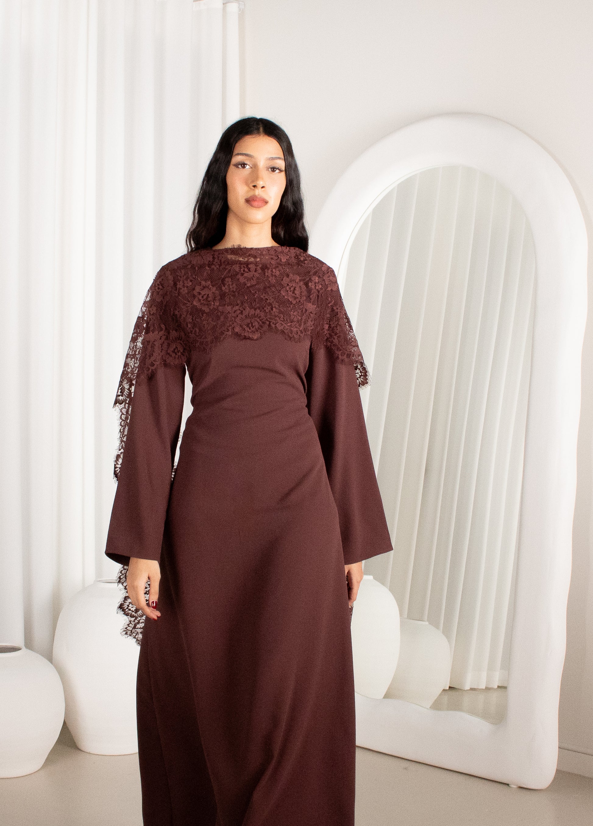 Woman wearing a brown dress with lace sleeves in a white room.