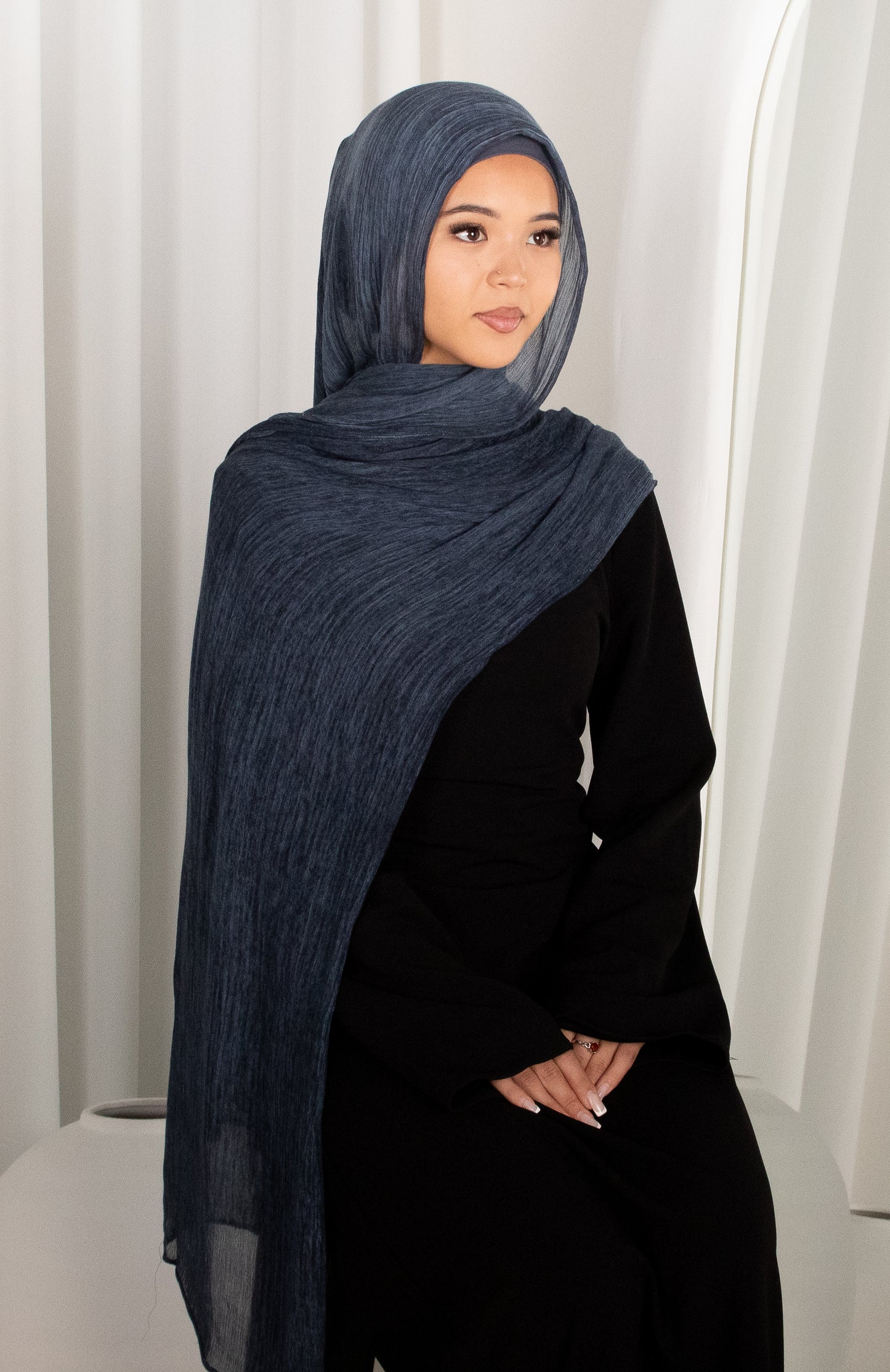 Woman wearing a dark blue hijab and black outfit against a white curtain background