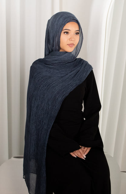 Woman wearing a dark blue hijab and black outfit against a white curtain background