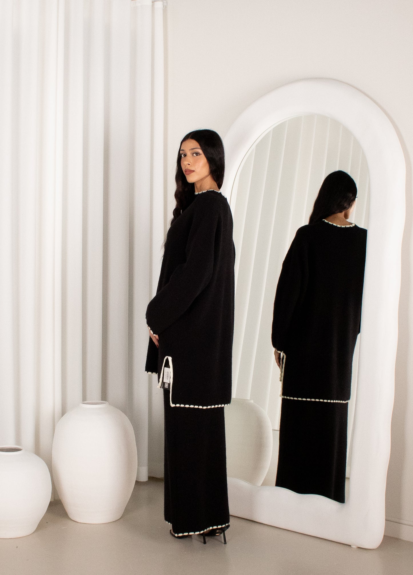 Woman in a black outfit standing in front of a white mirror with a white curtain background.
