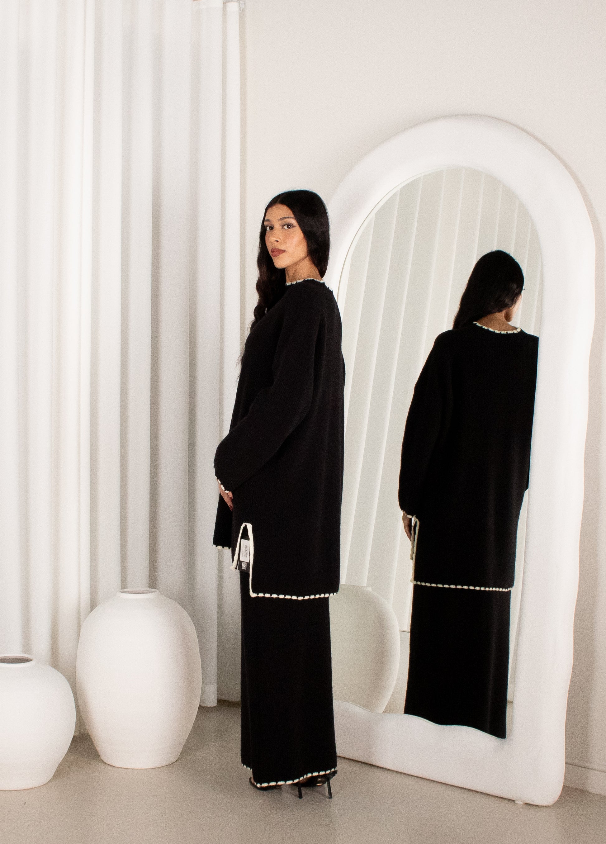 Woman in a black outfit standing in front of a white mirror with a white curtain background.