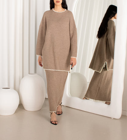 Woman in a brown outfit standing in front of a mirror with white curtains and a white vase in the background.