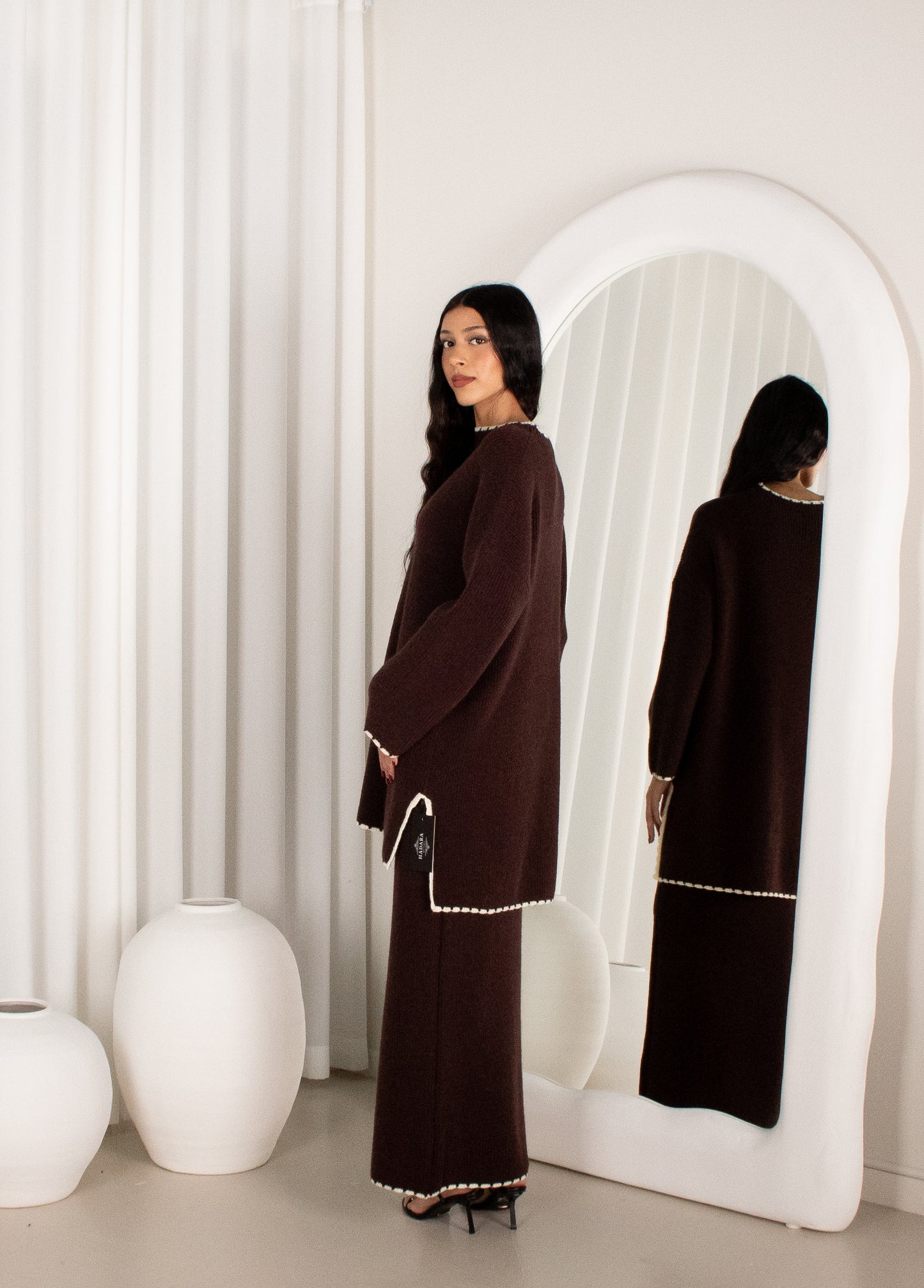 Woman in a brown dress standing in front of a white mirror with a white curtain background.