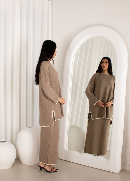 Woman in a beige outfit standing in front of a large mirror in a minimalistic room.