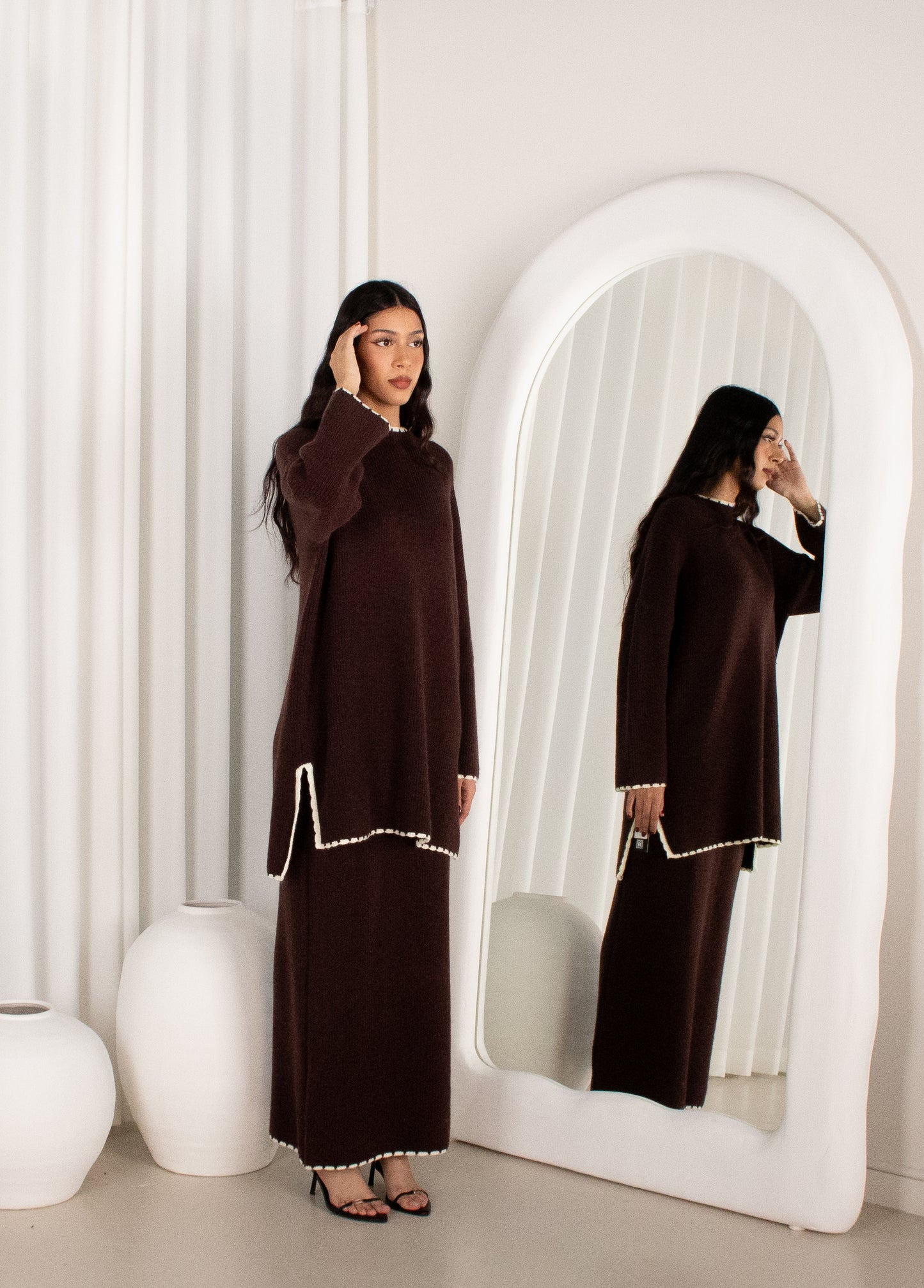 Woman in a brown dress standing in front of a large mirror in a minimalistic room.