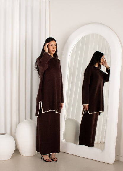 Woman in a brown dress standing in front of a large mirror in a minimalistic room.