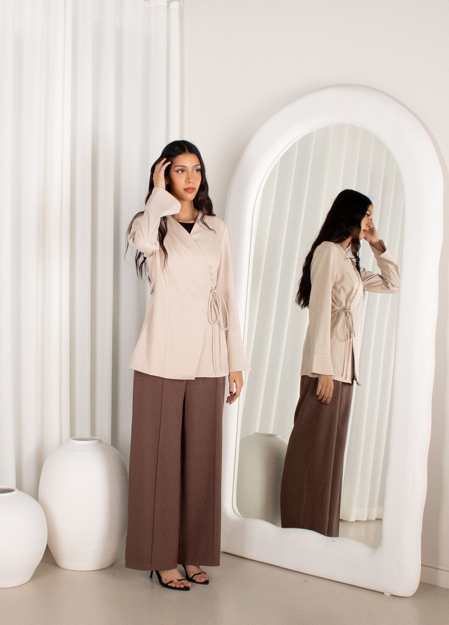 Woman adjusting her hair in front of a large mirror wearing a beige top and brown pants.