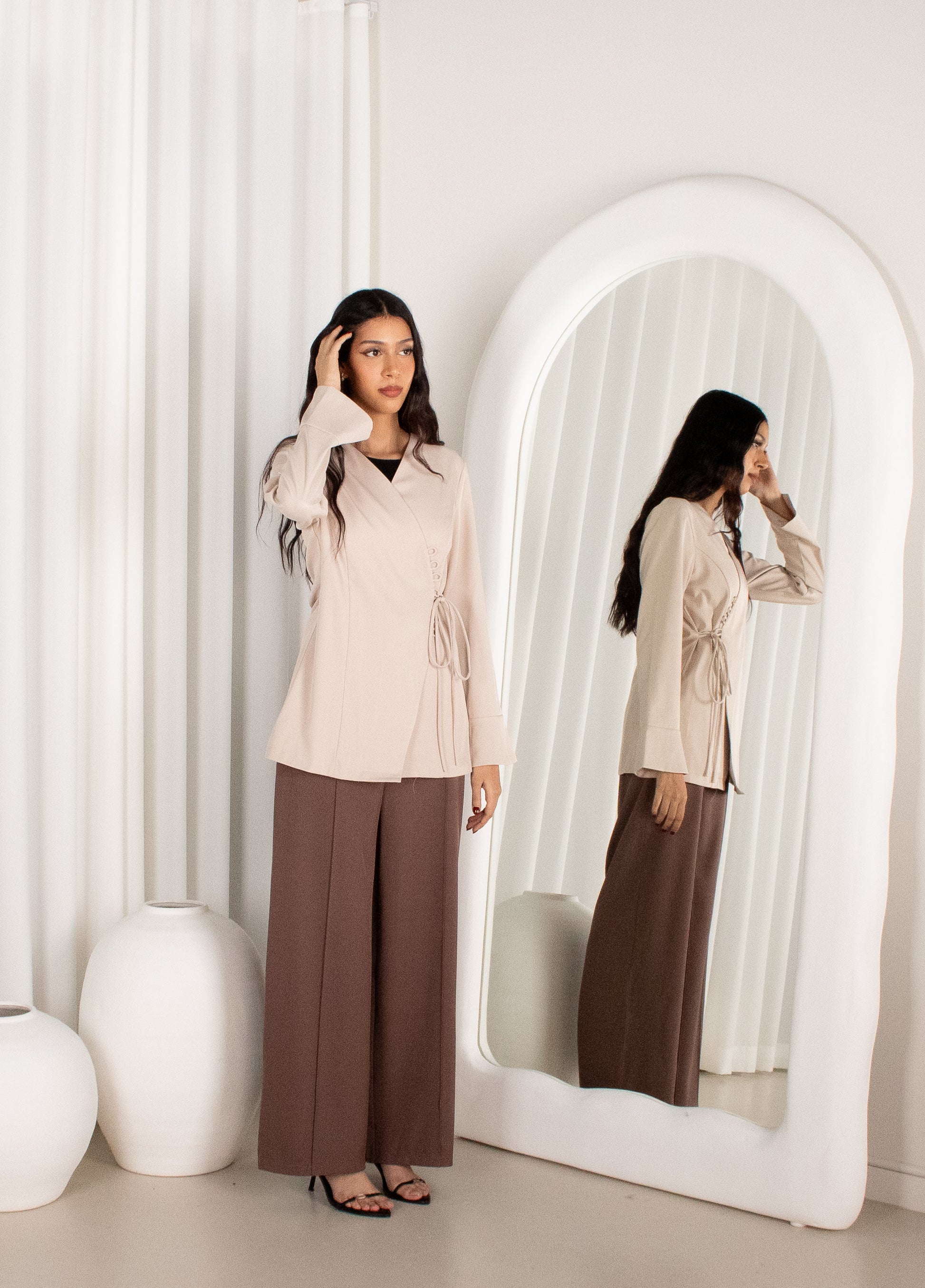 Woman adjusting her hair in front of a large mirror wearing a beige top and brown pants.
