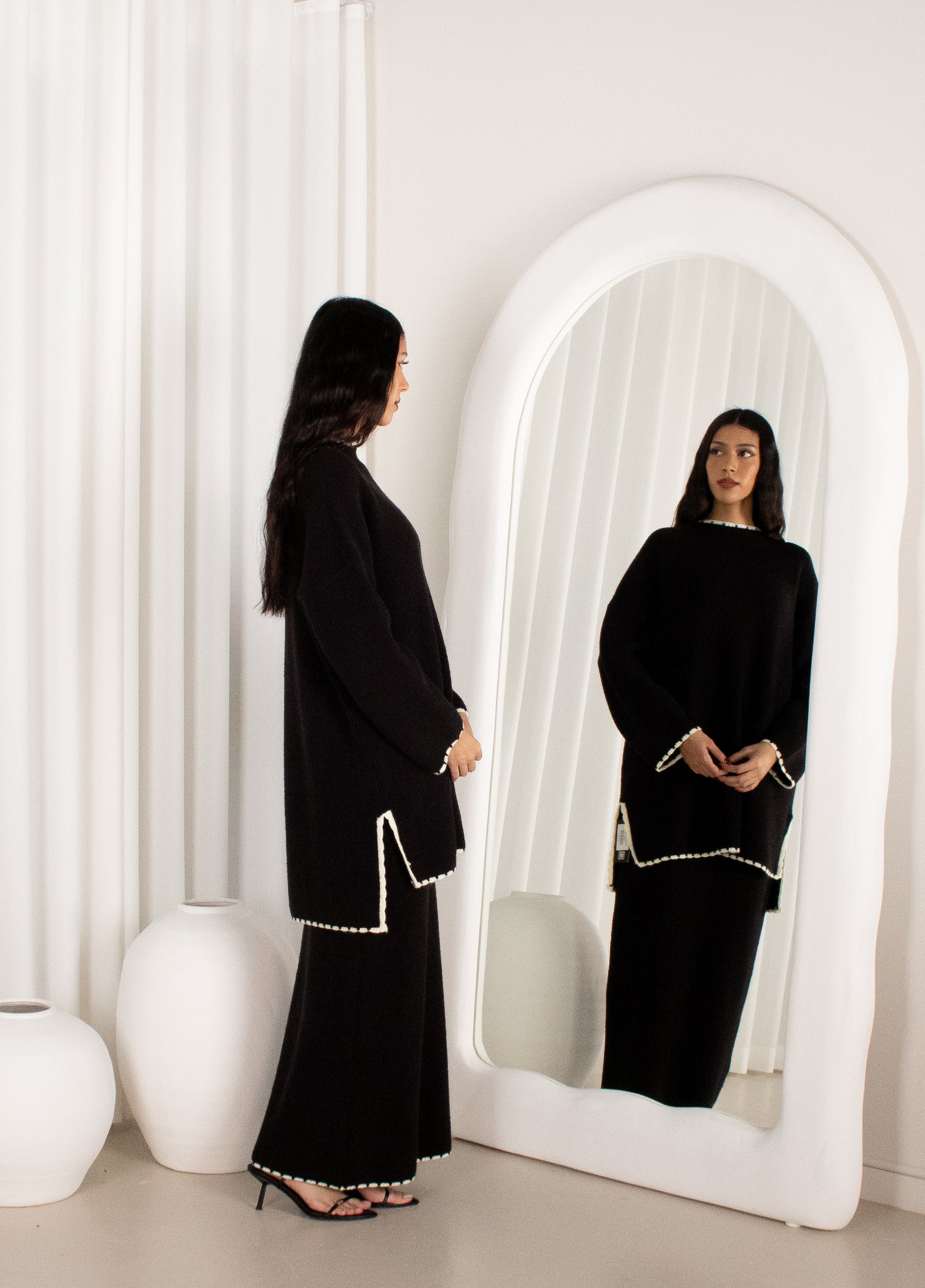 Woman in a black outfit standing in front of a white mirror in a minimalistic room.
