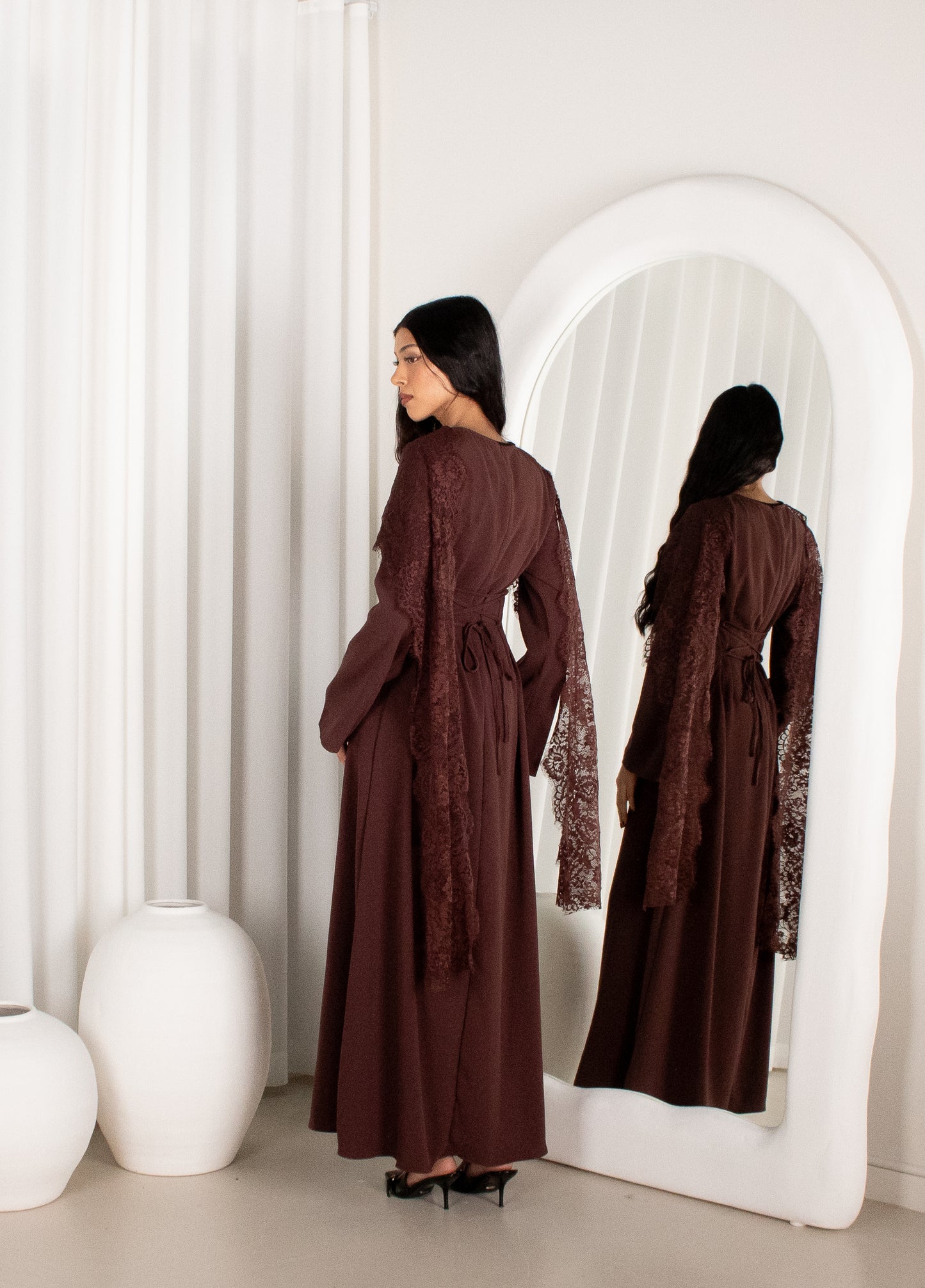 Woman in a brown dress standing in front of a mirror in a minimalistic room.