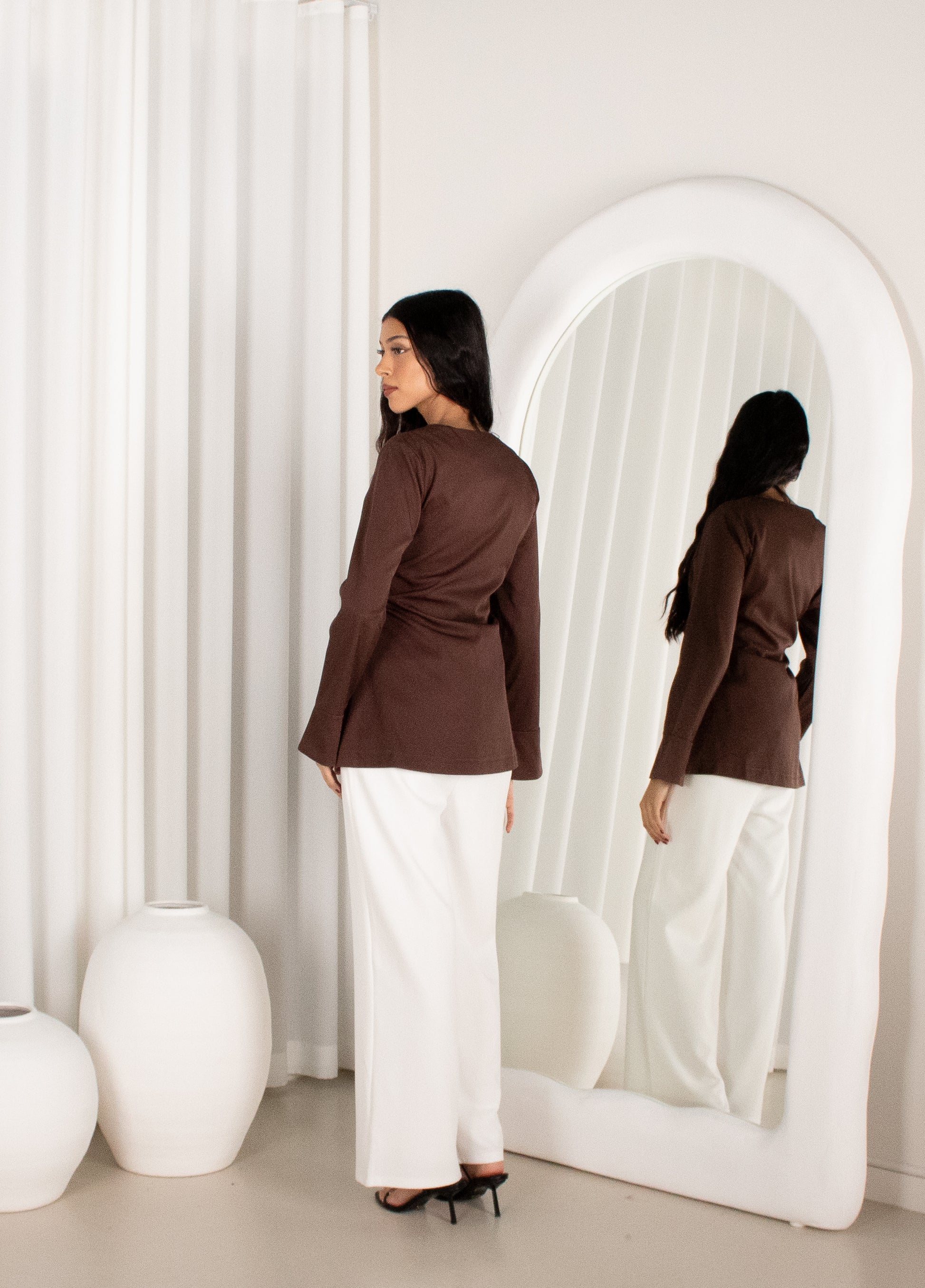 Woman in a brown top and white pants standing in front of a large mirror in a minimalistic room.