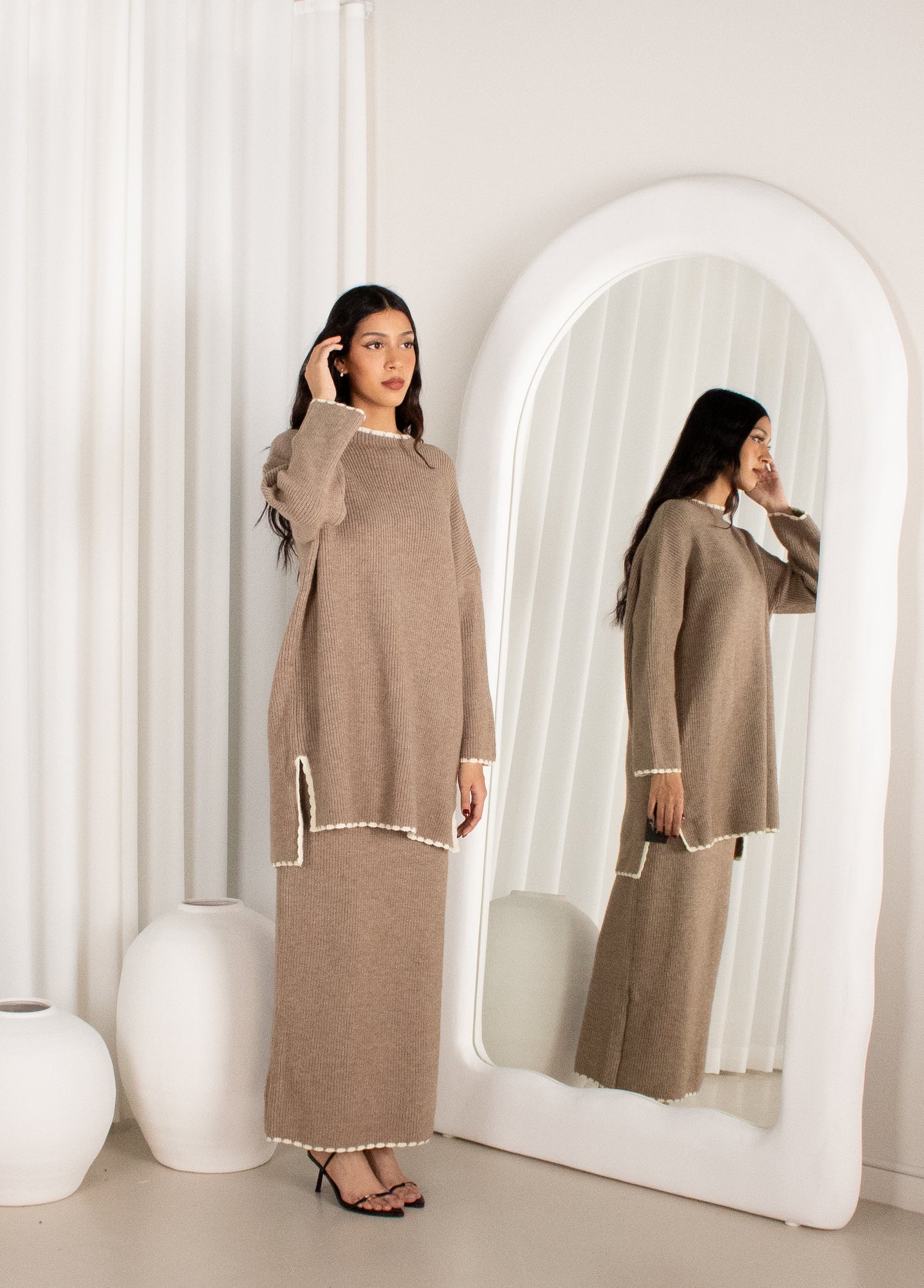 Woman in a beige outfit standing in front of a mirror in a minimalistic room.