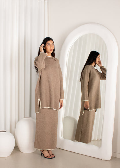Woman in a beige outfit standing in front of a mirror in a minimalistic room.