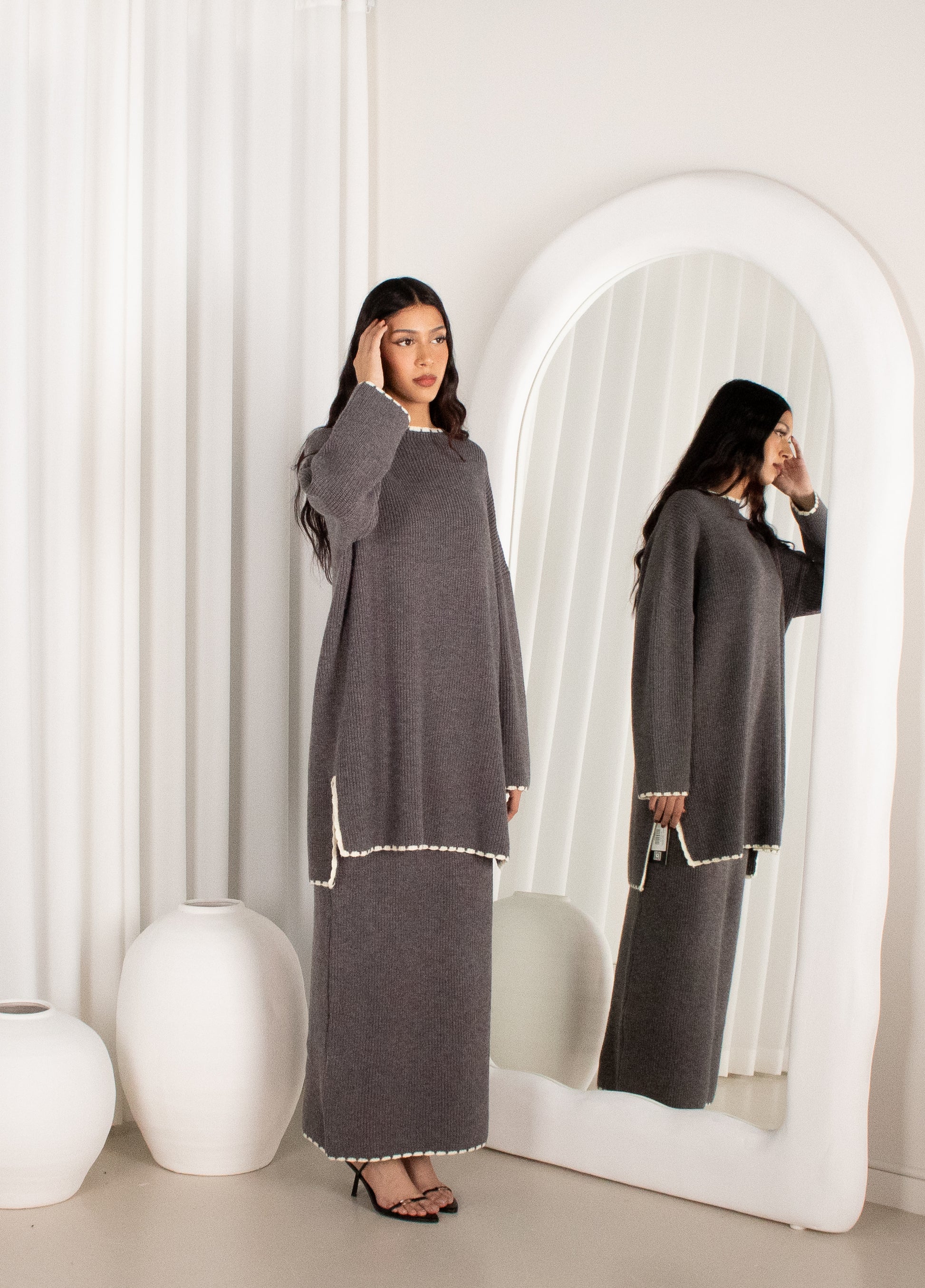 Woman in a gray dress standing in front of a mirror in a minimalistic room.
