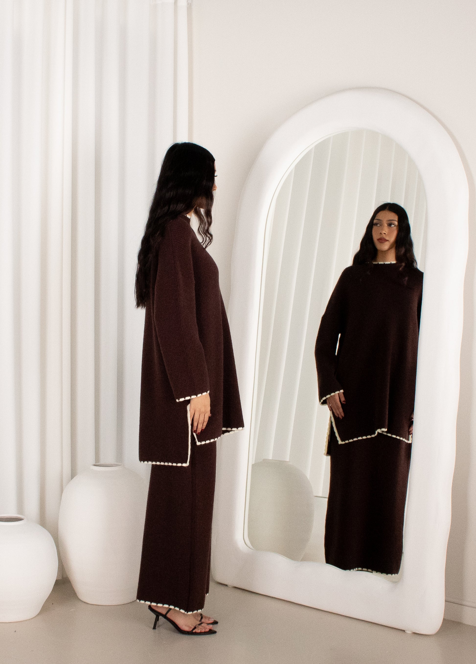 Woman in a dark brown outfit standing in front of a white mirror.