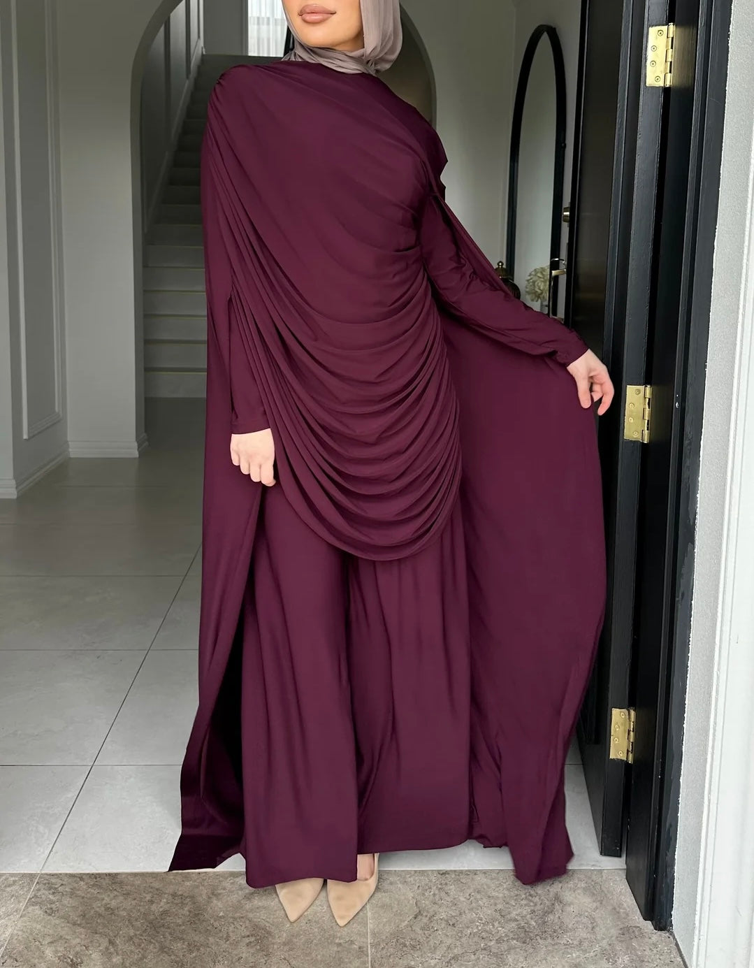 Woman wearing a burgundy long-sleeve dress with a draped front, standing in a doorway.