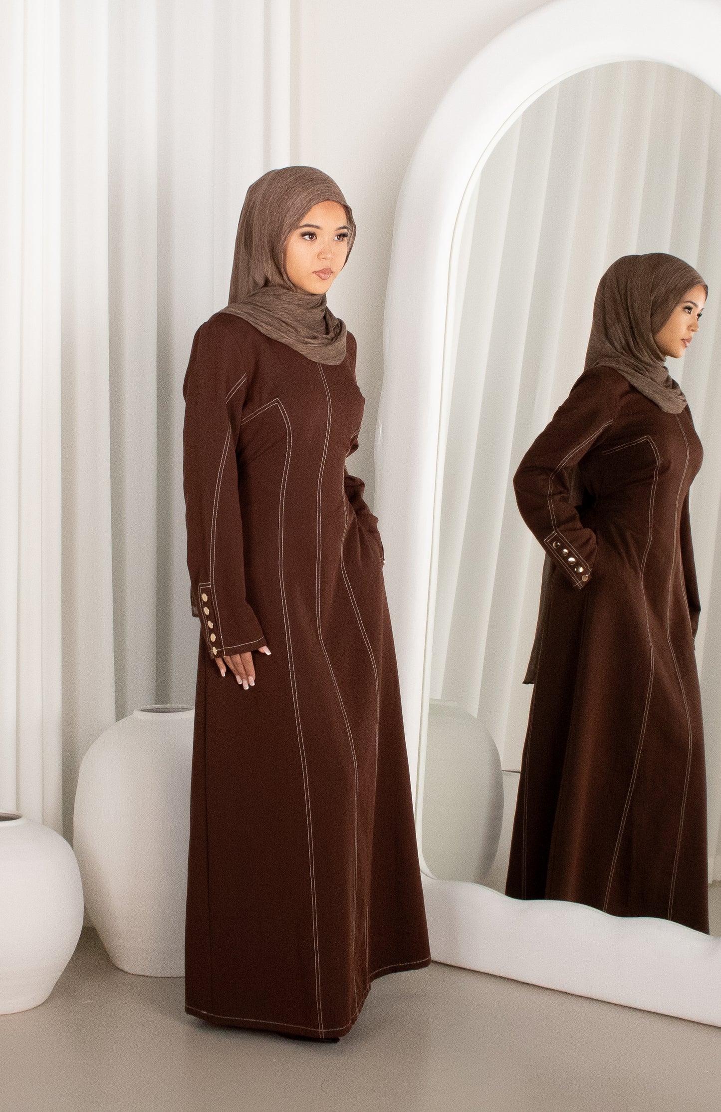 Woman in a brown dress and hijab standing in front of a mirror.