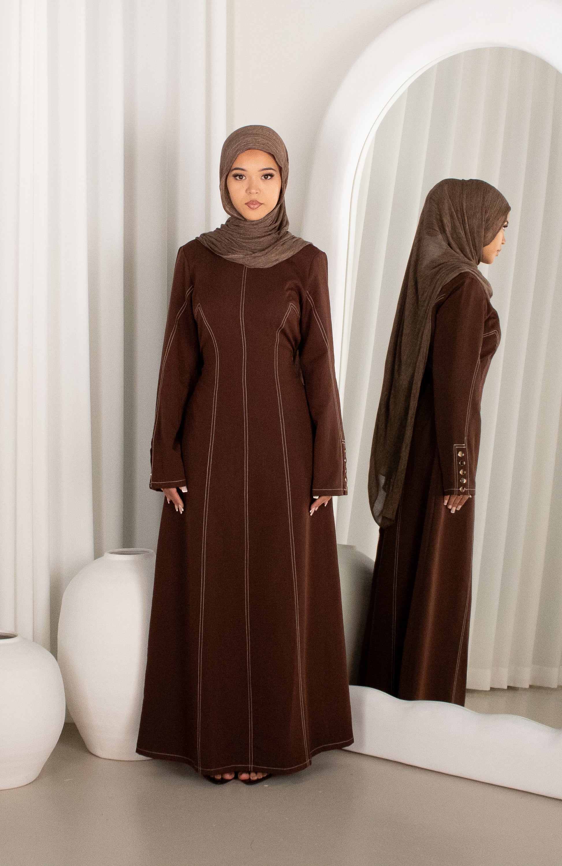 Woman in a brown abaya standing in front of a mirror with a white curtain background
