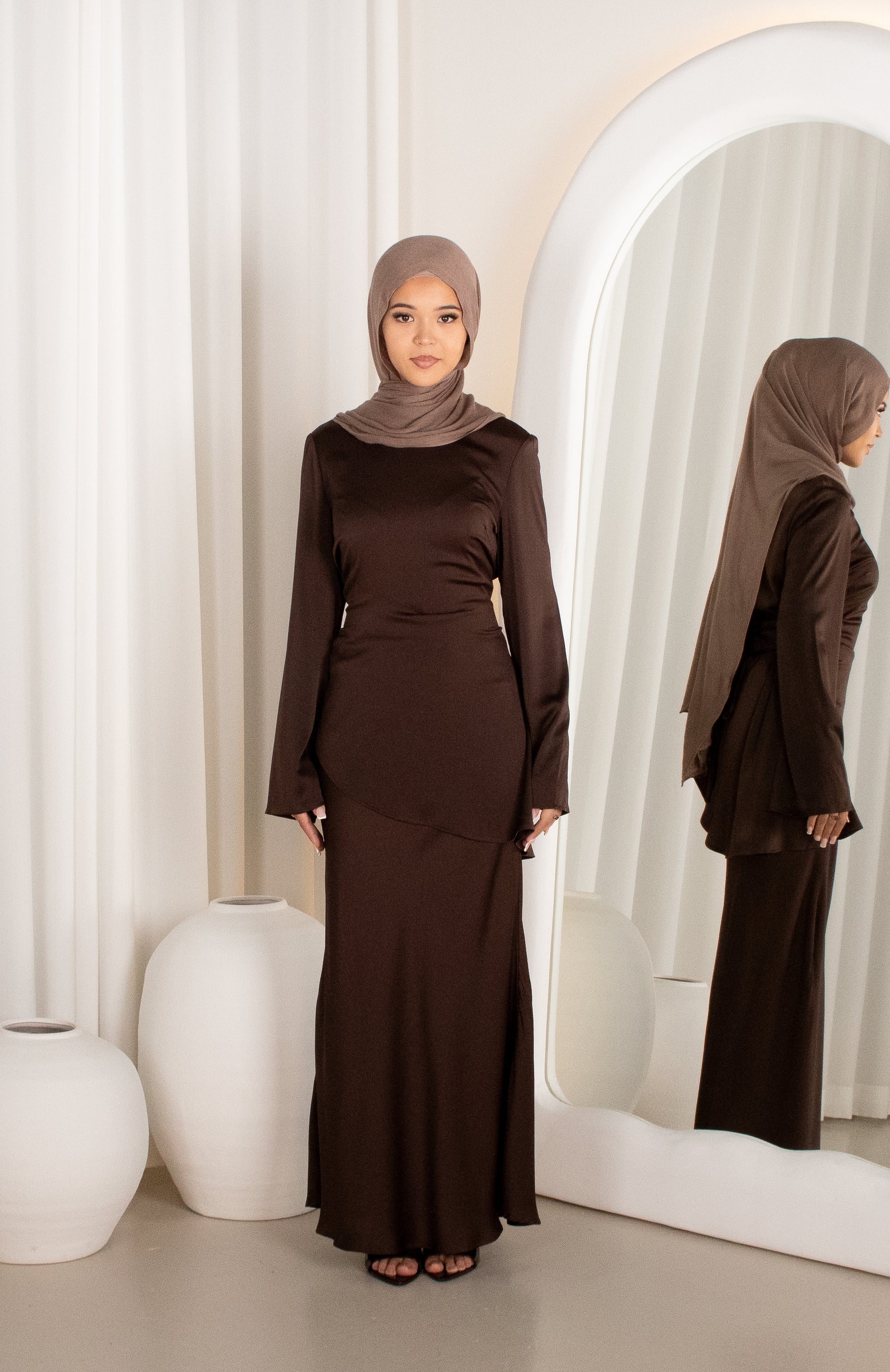 Woman in a brown outfit standing in front of a mirror with white curtains.