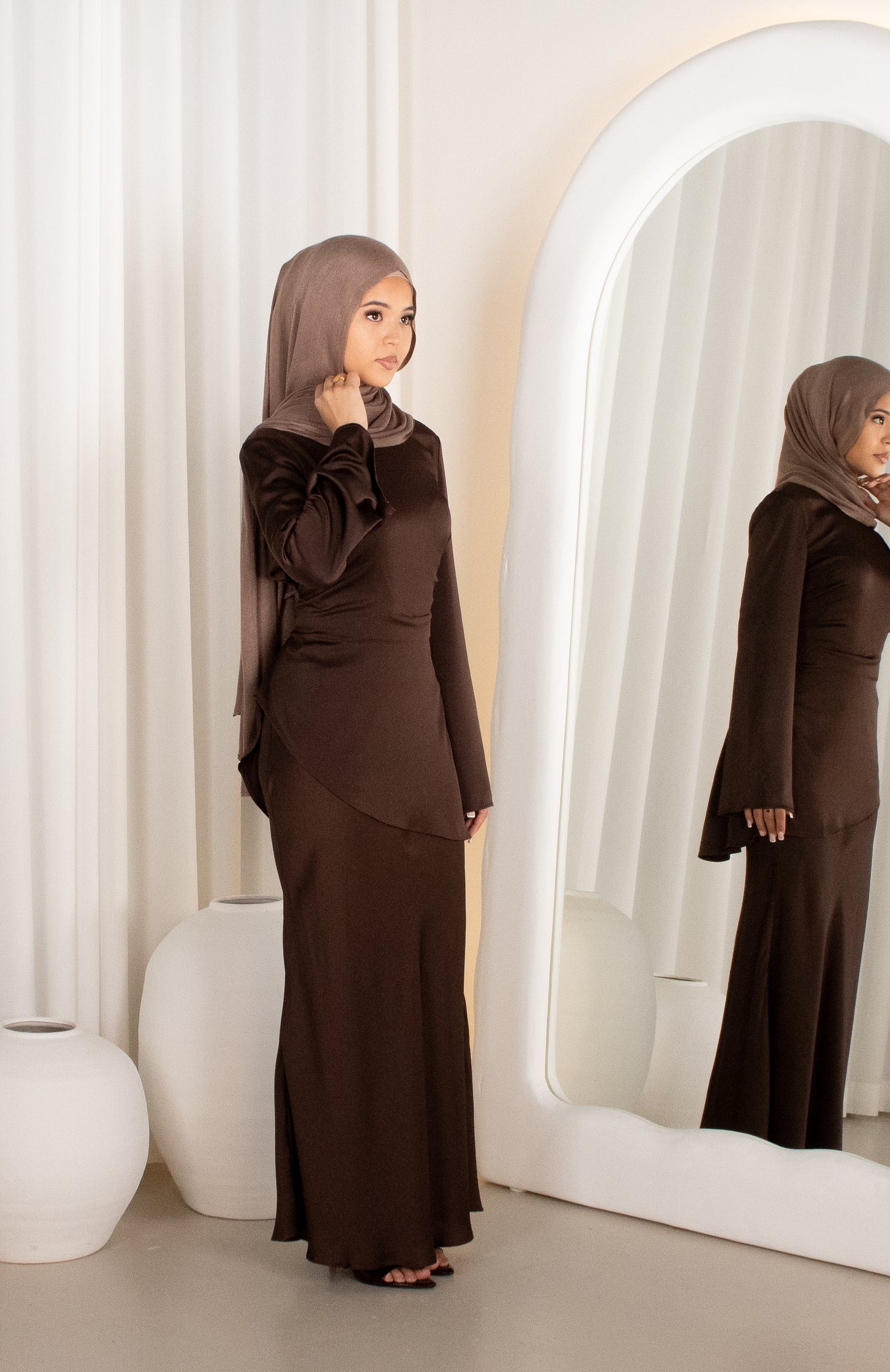 Woman in a brown outfit standing in front of a mirror with a white curtain background