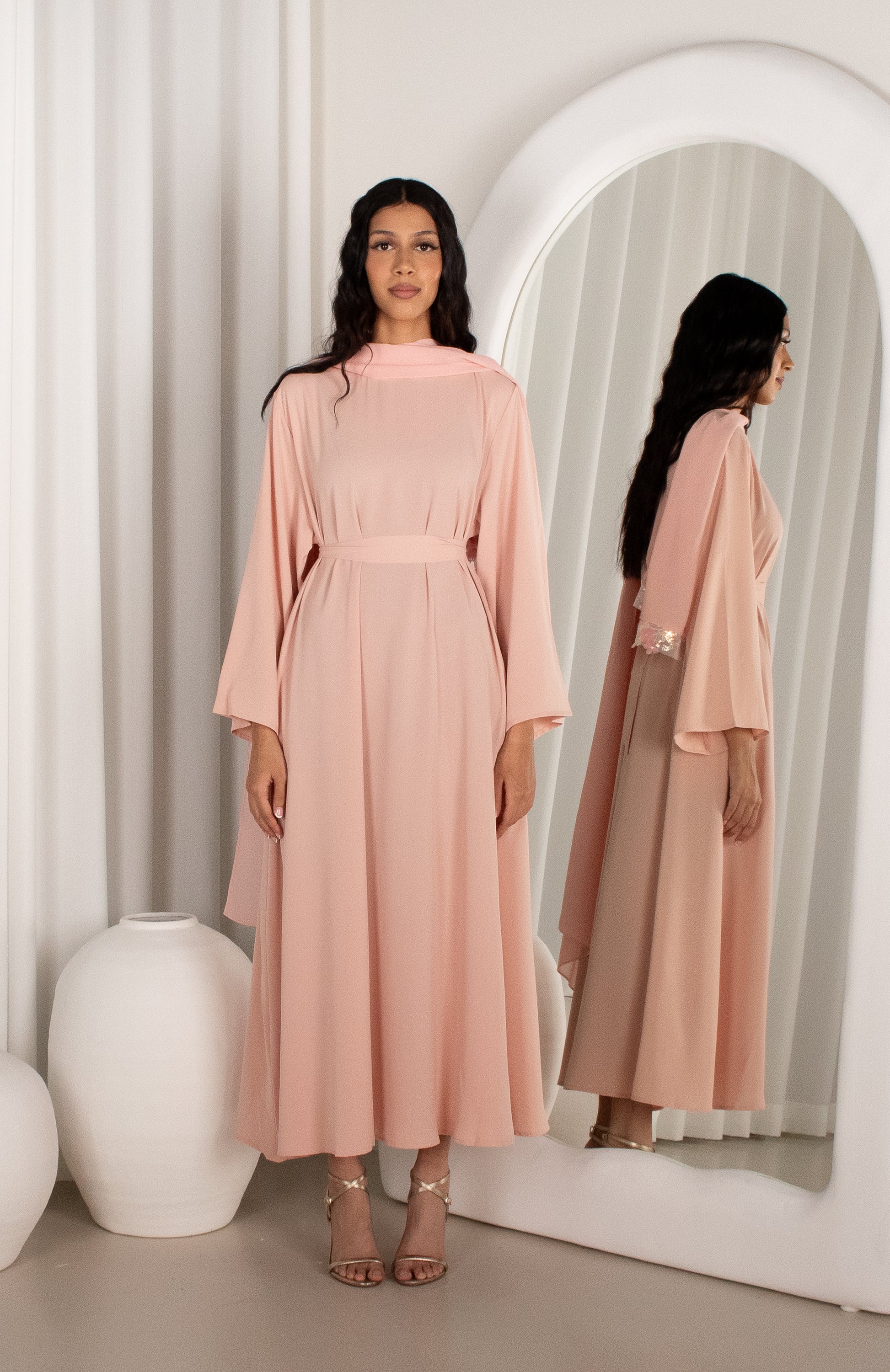 Woman in a pink dress standing in front of a mirror with a white curtain background